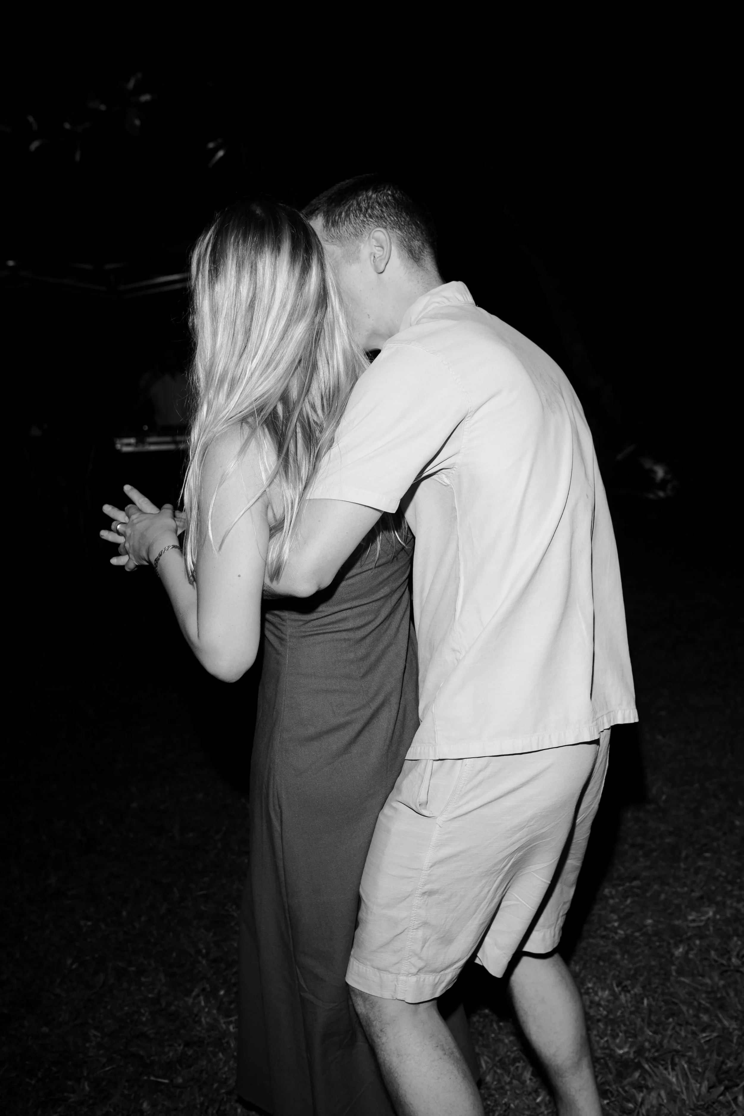 A black-and-white photo of a young couple dancing closely outdoors at night, with their faces touching and the woman's long hair down.