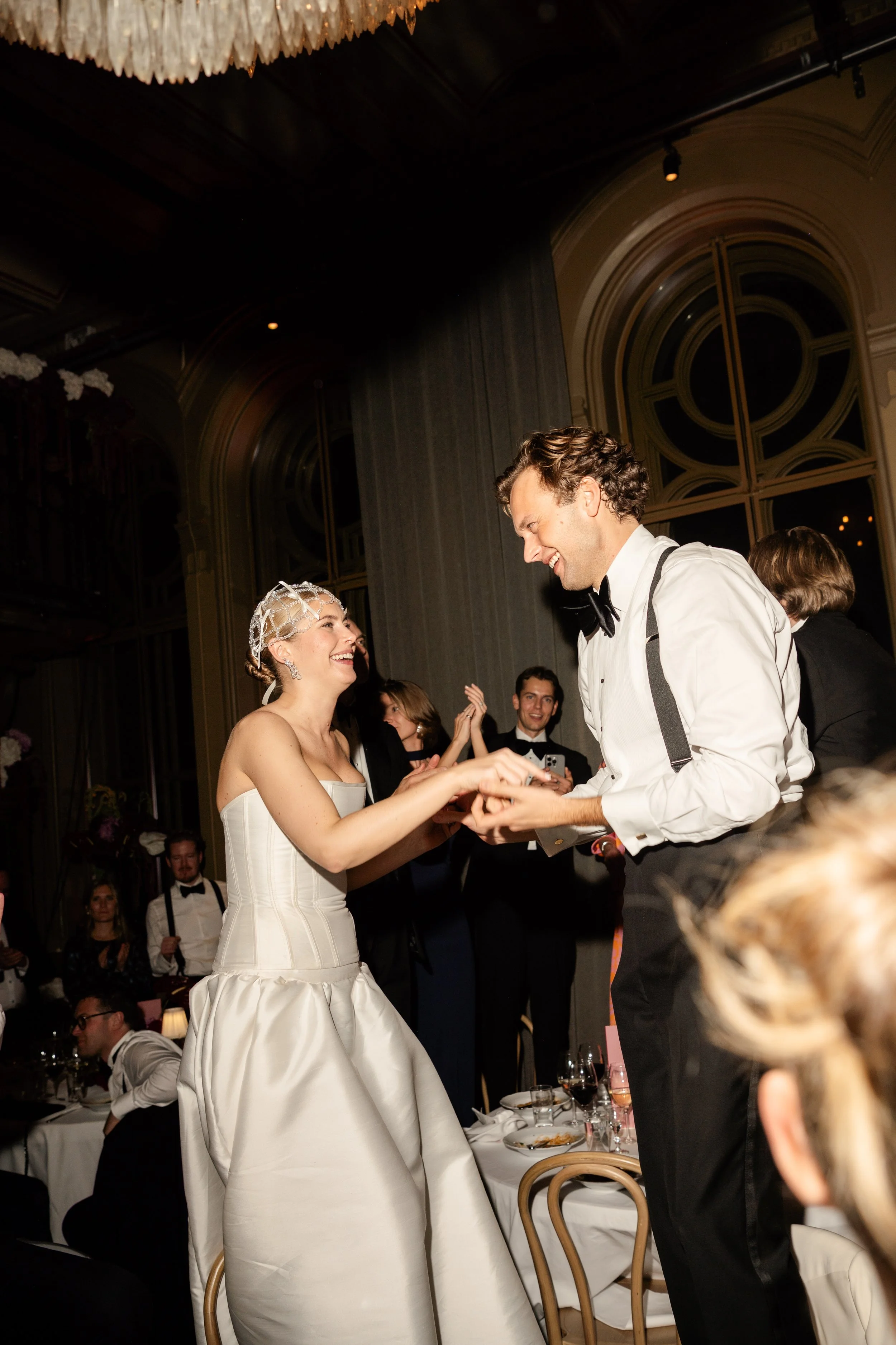 A bride and groom are smiling and dancing together at their wedding reception, surrounded by friends and guests in an elegant, dimly lit banquet hall.
