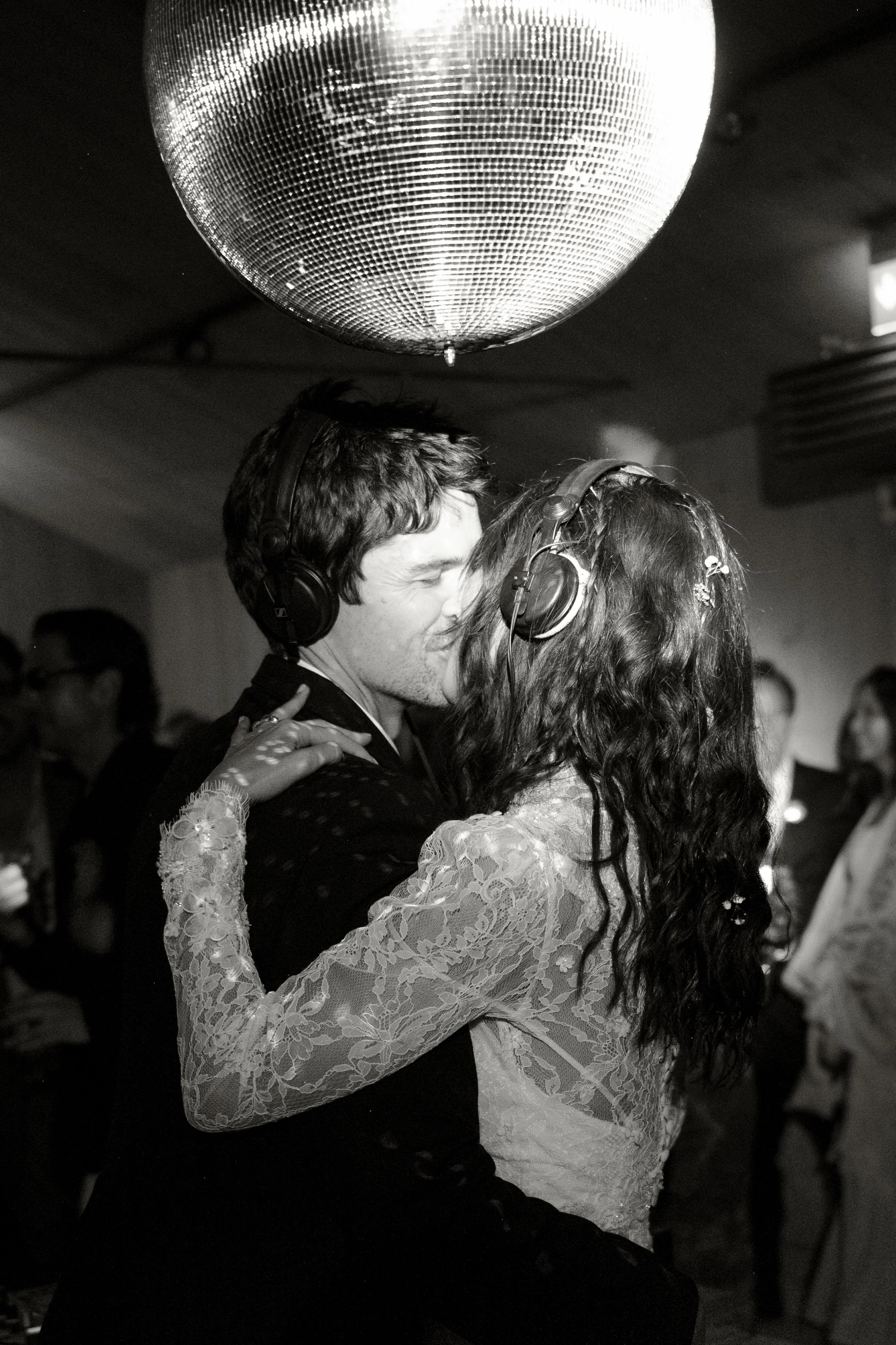 A black and white photo of a couple dancing closely at a party, both wearing headphones, with a disco ball overhead and other people in the background.