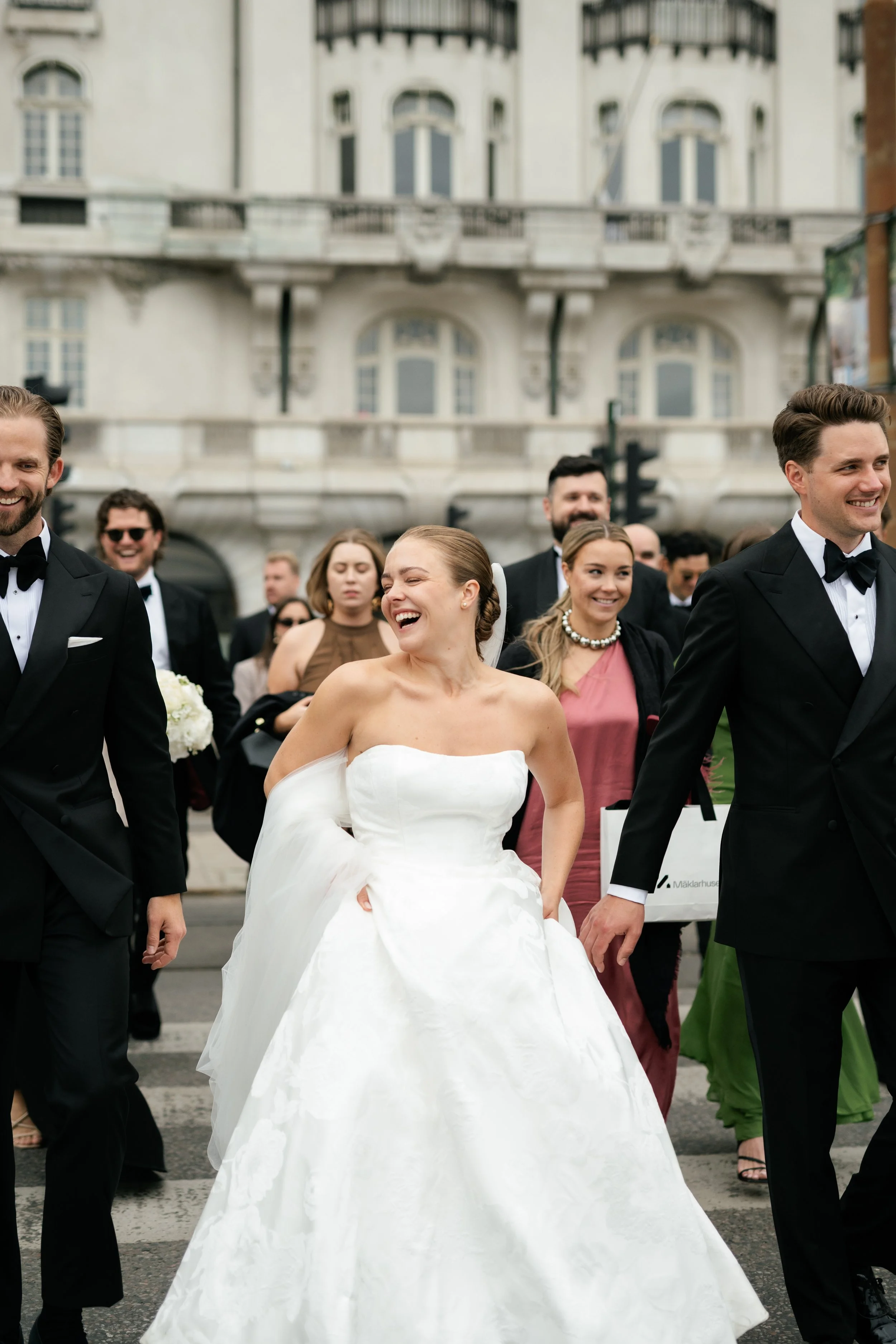 People dressed in formal attire walking on a city street during a wedding celebration, with a woman in a white wedding gown smiling and holding her dress.