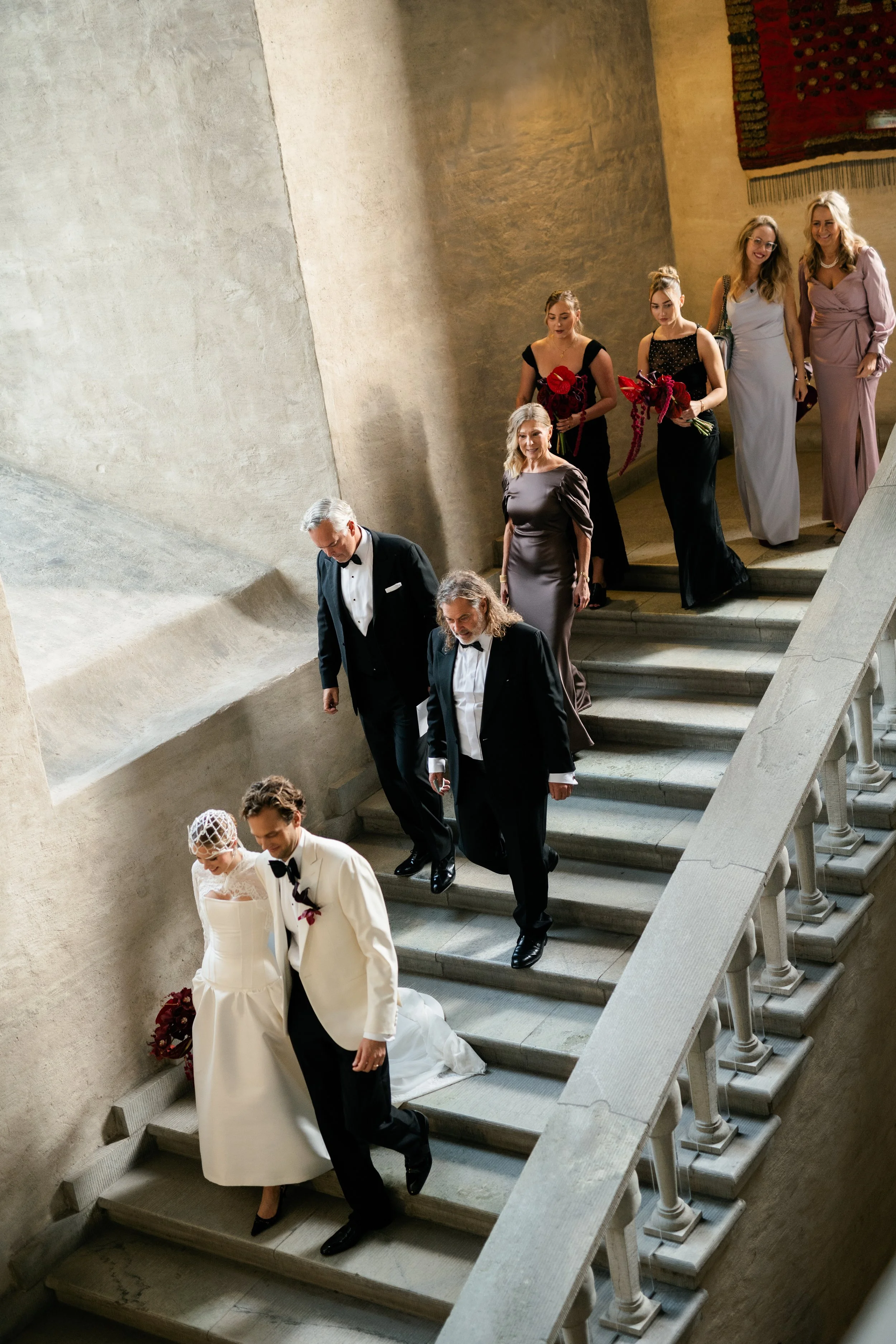 A wedding celebration with the bride and groom descending a stone staircase, accompanied by family and friends, in an elegant indoor setting.