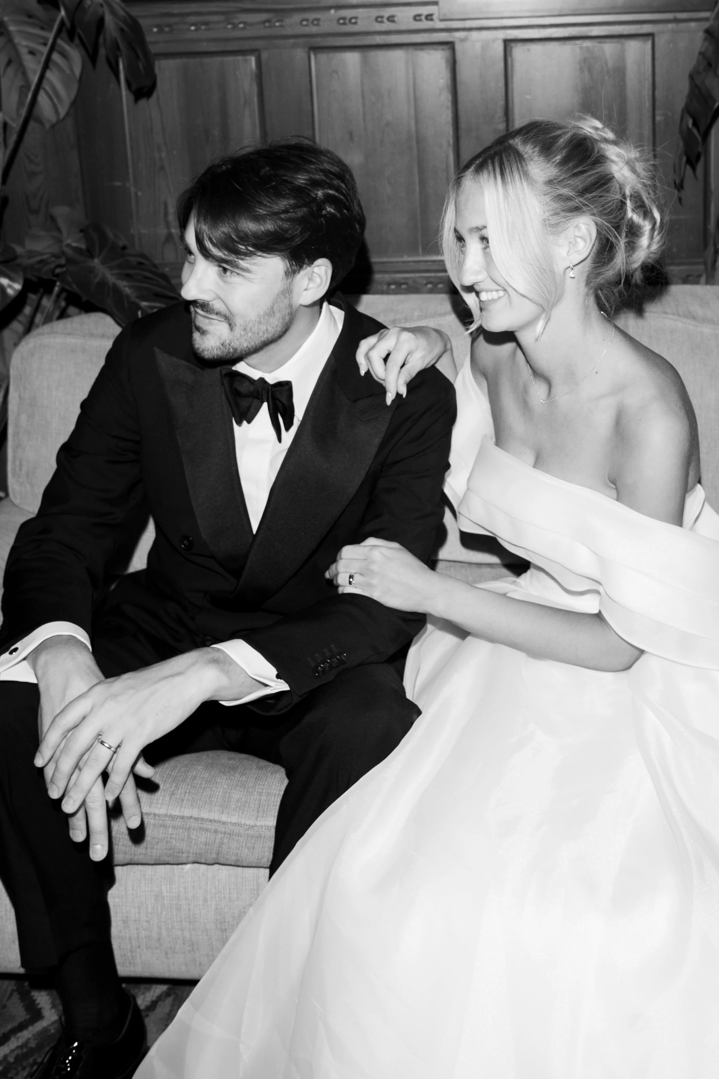 A black and white photo of a couple dressed in formal wedding attire sitting on a sofa, smiling.