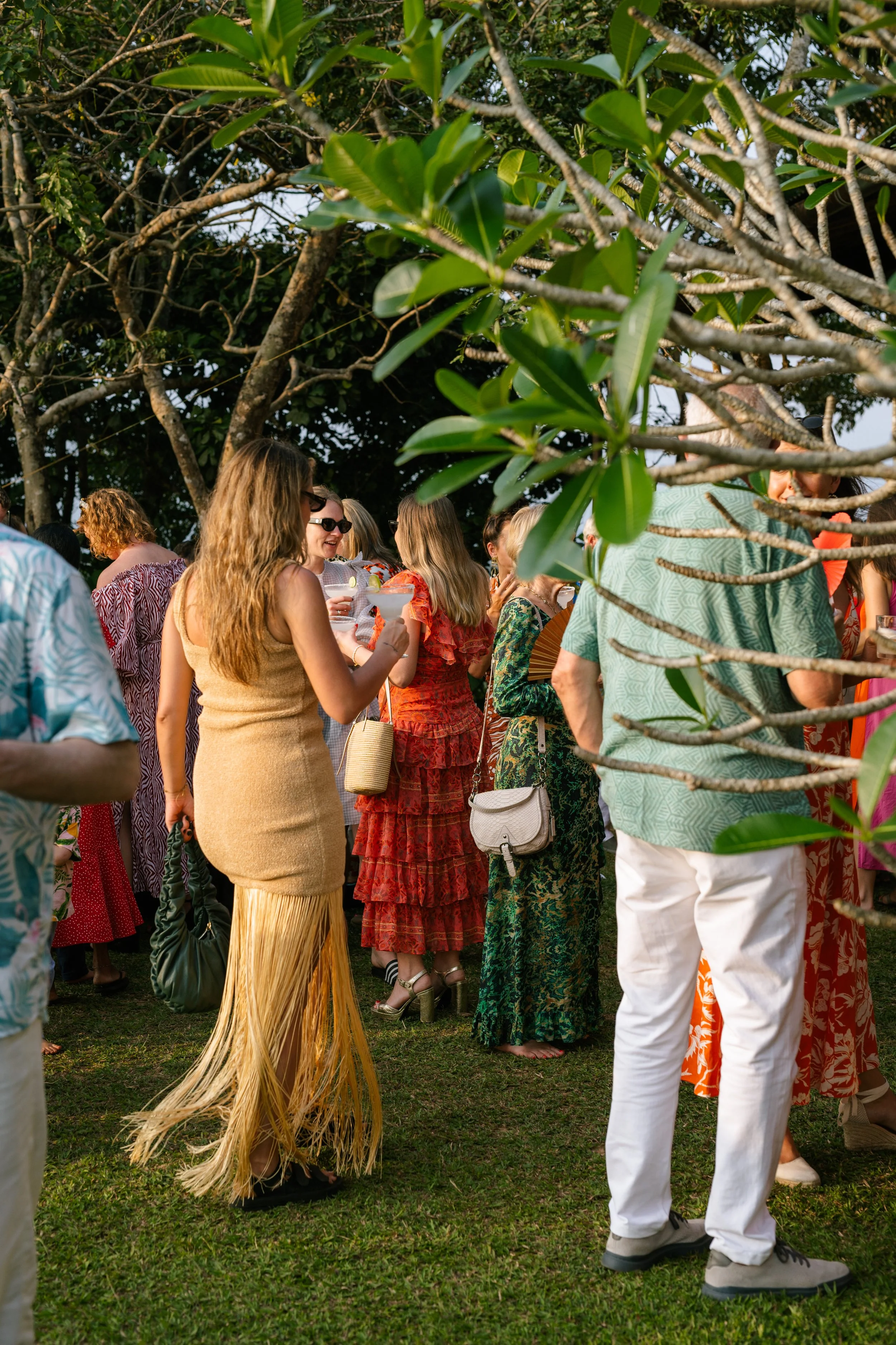 People gathered outdoors under trees at a social event, some holding drinks and wearing colorful, summery dresses.