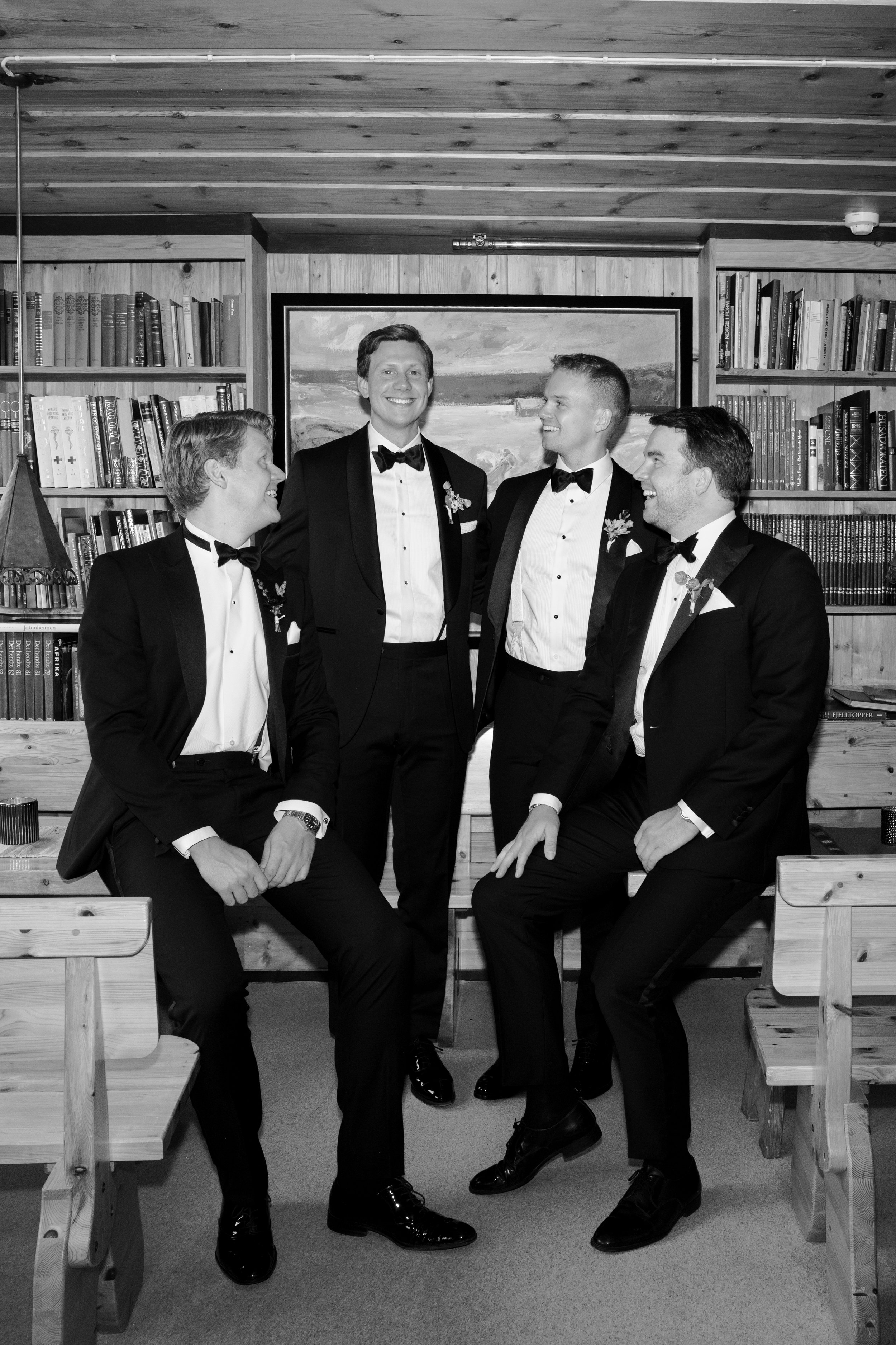 Four men in tuxedos in a room with wooden walls and bookshelves, engaged in conversation and smiling.