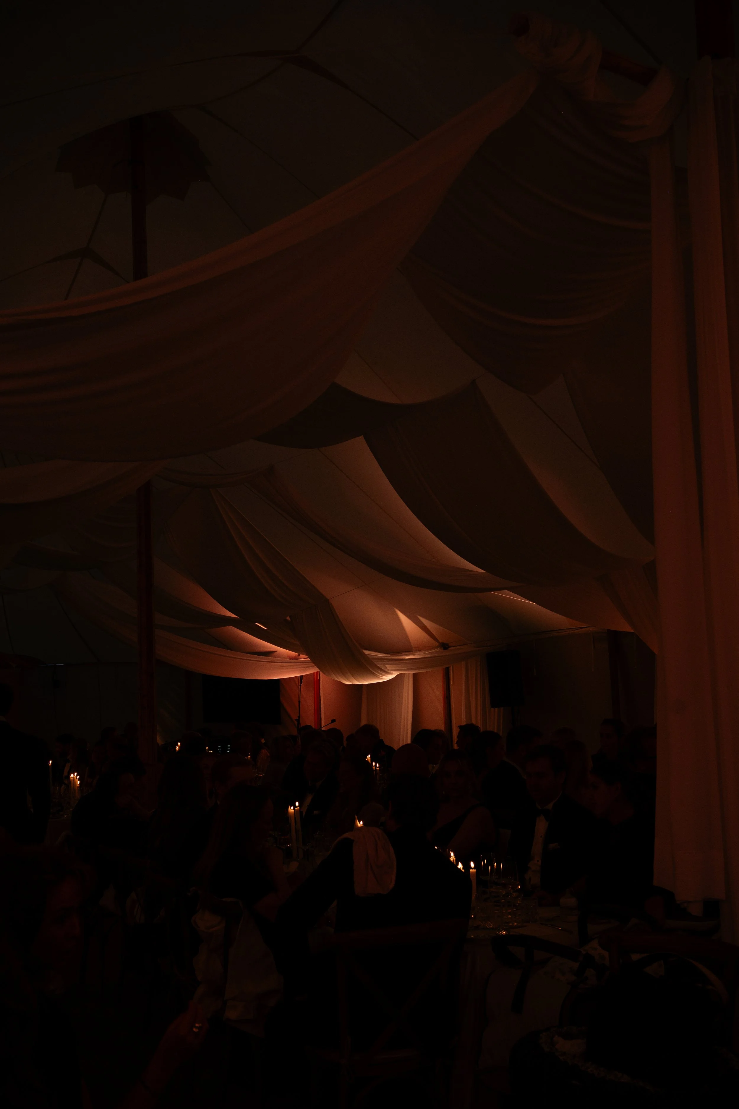 Dimly lit banquet hall with draped fabric ceiling and candlelit tables, filled with seated guests in formal attire.
