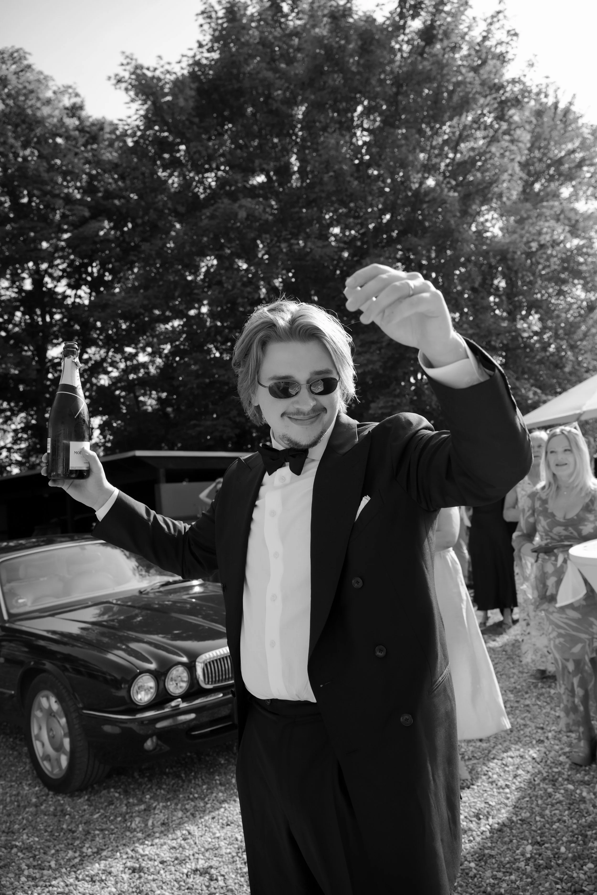 A man dressed in a tuxedo with a bow tie and sunglasses, holding a bottle of champagne in one hand, smiling and raising his other arm at an outdoor gathering with people and a vintage car in the background.