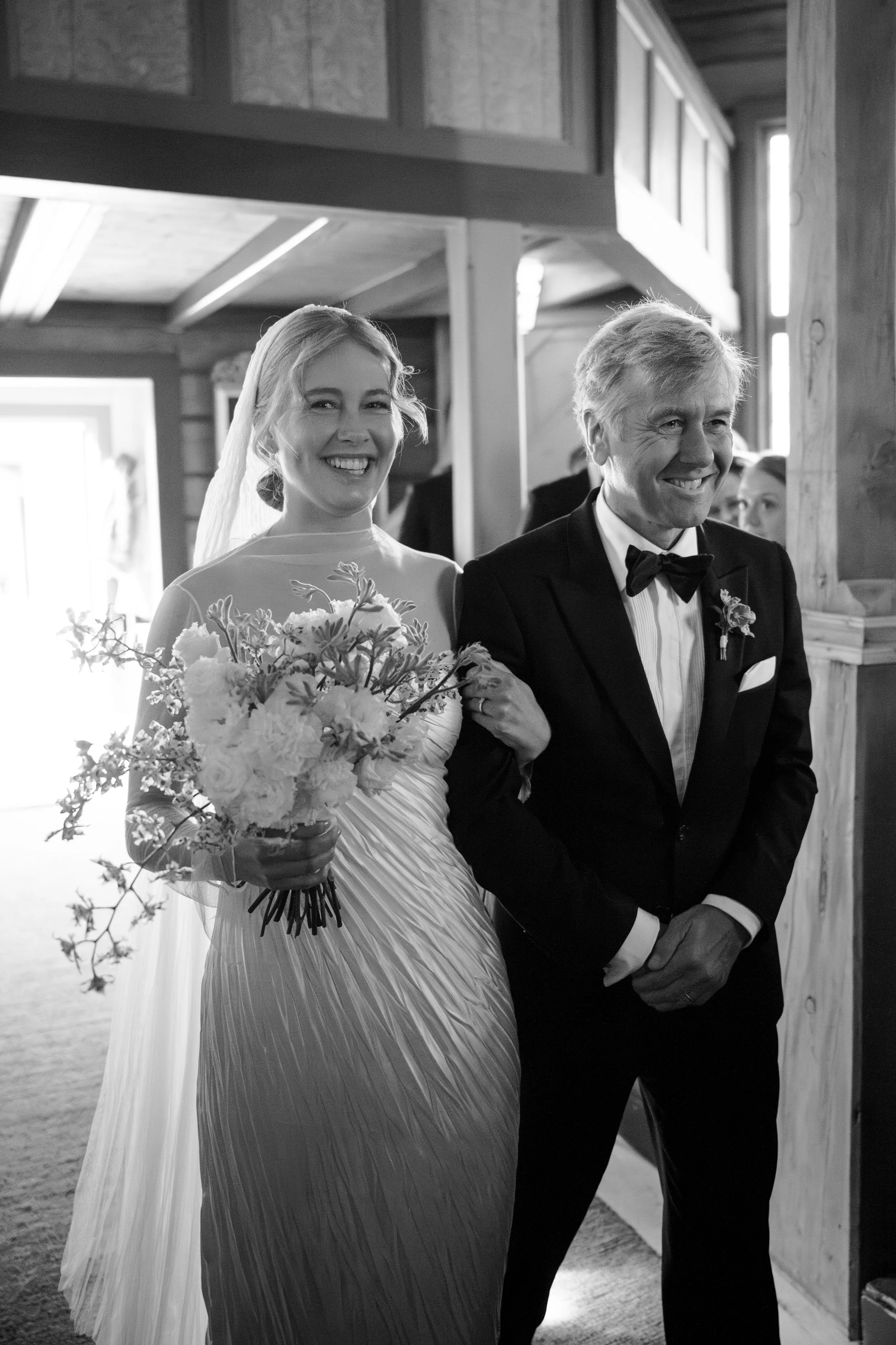 A bride in a wedding dress holding a bouquet, walking down the aisle with an older man in a tuxedo, at a wedding ceremony in a wooden venue.