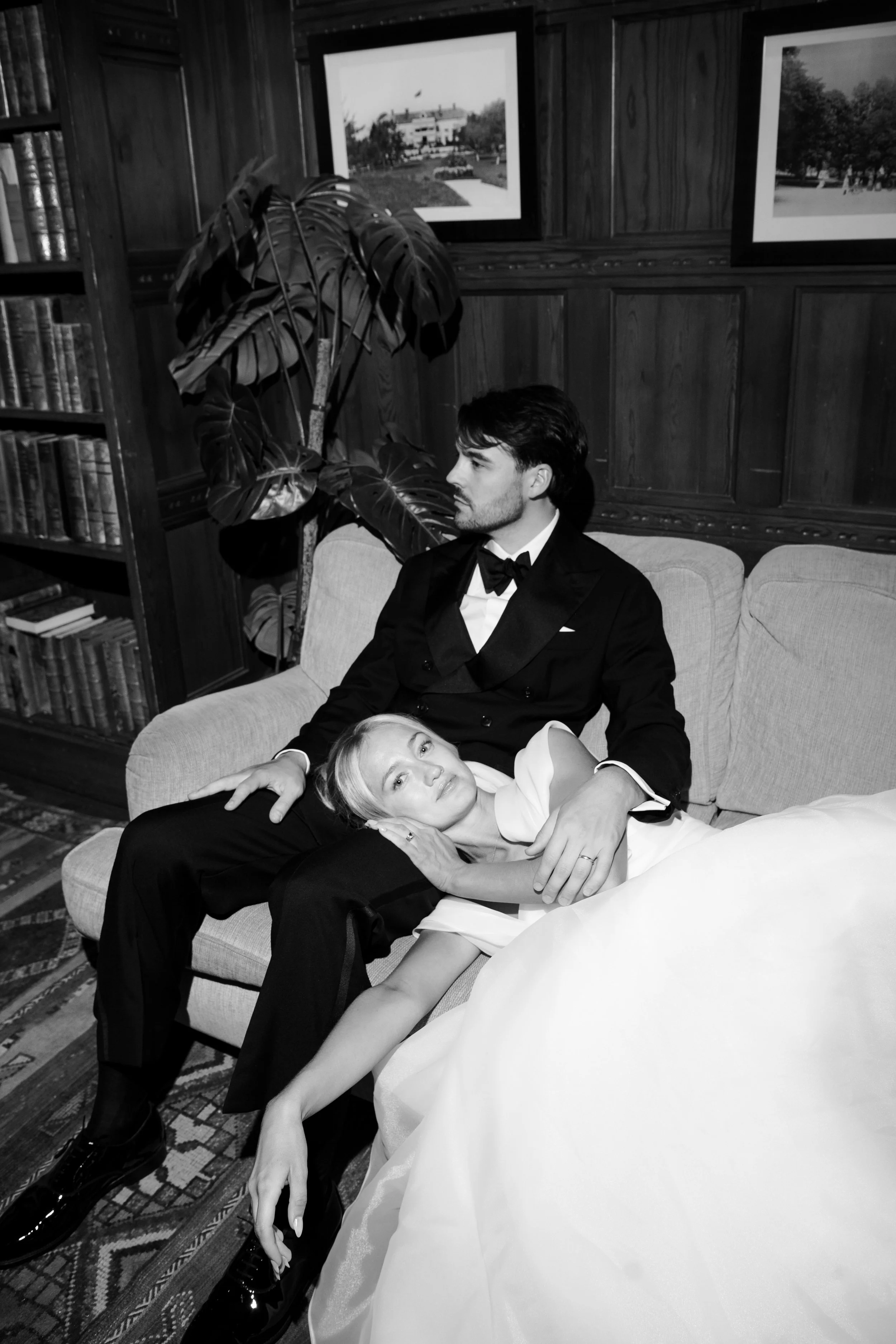 A black and white photo of a man and woman, dressed formally, sitting on a sofa in a richly decorated room. The woman is lying with her head on the man's lap, and they are holding hands. The room features wood-paneled walls, framed photographs, a boo