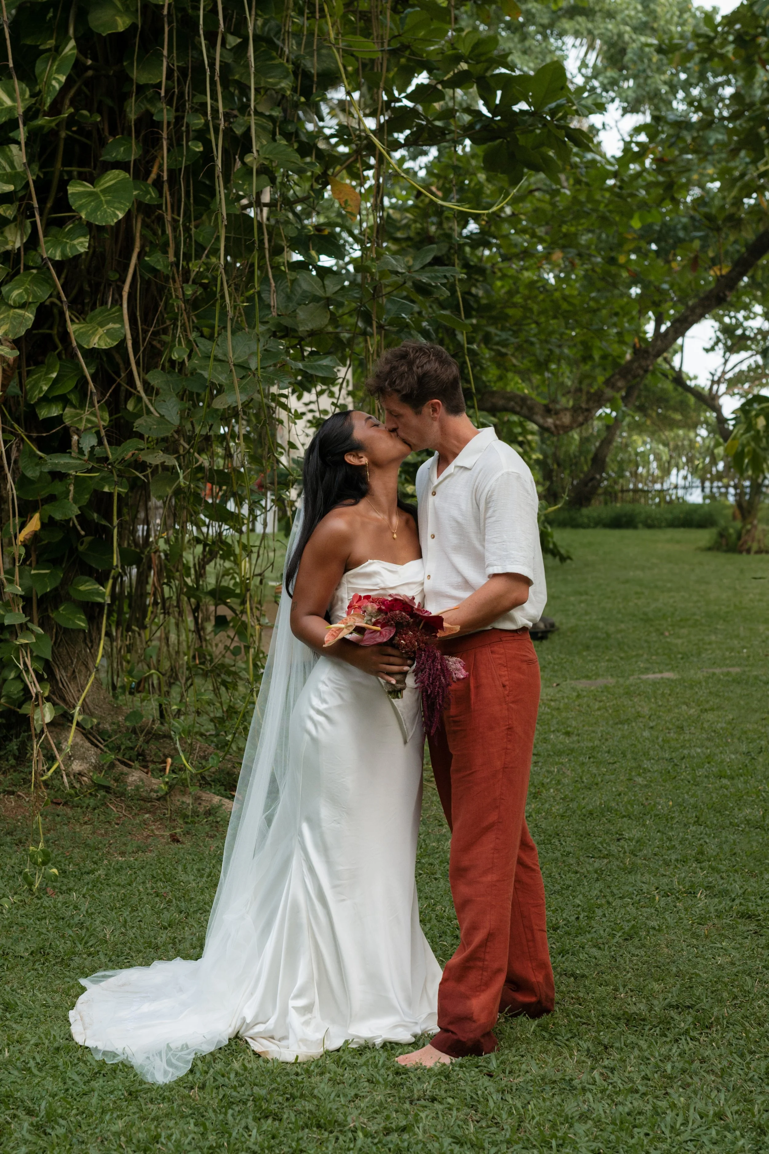 A bride and groom kissing outdoors, with the bride holding a bouquet, under green trees and grass.