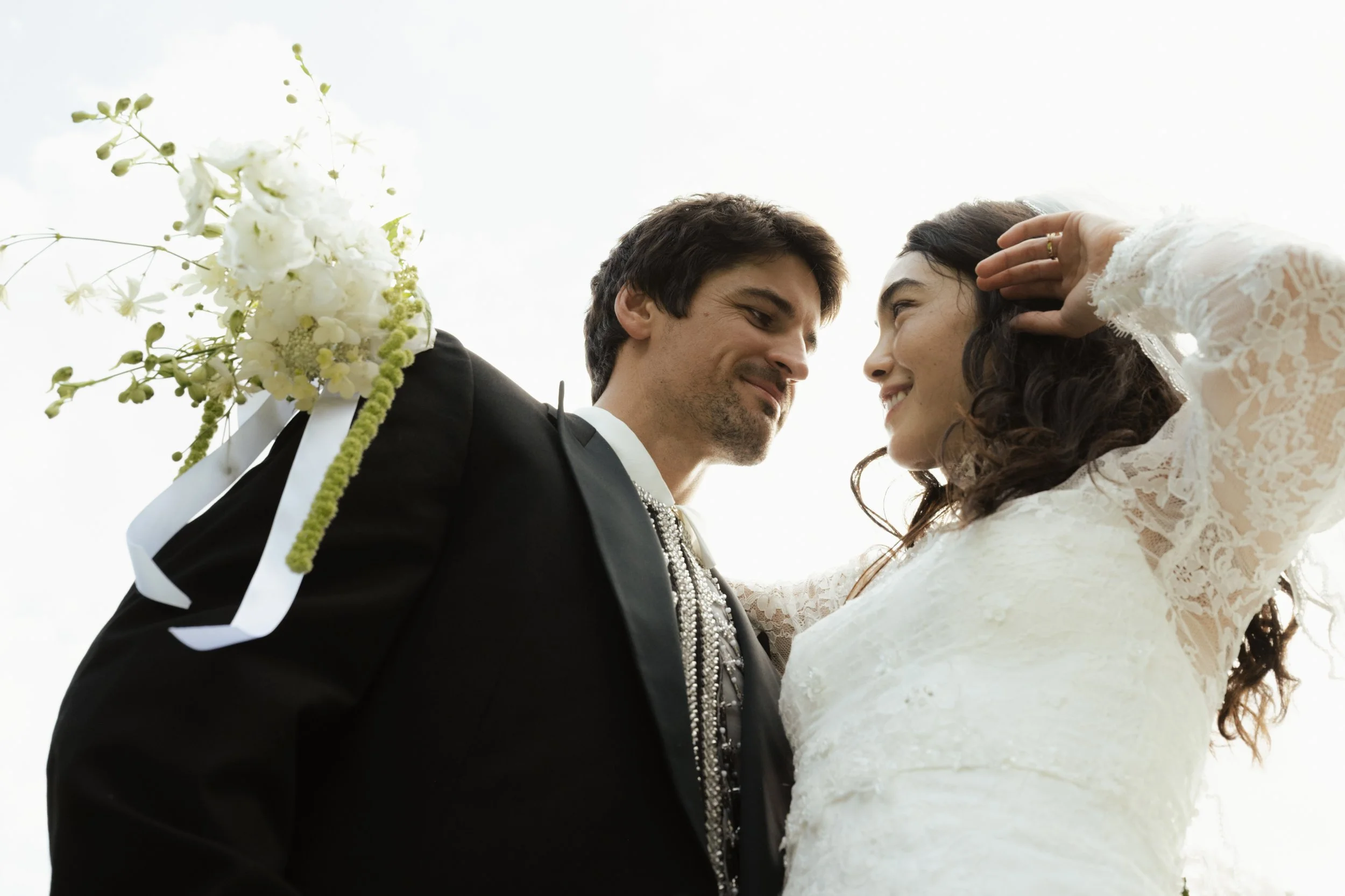 A happy couple on their wedding day looking at each other, with the bride touching her hair and the groom holding a bouquet of white flowers.