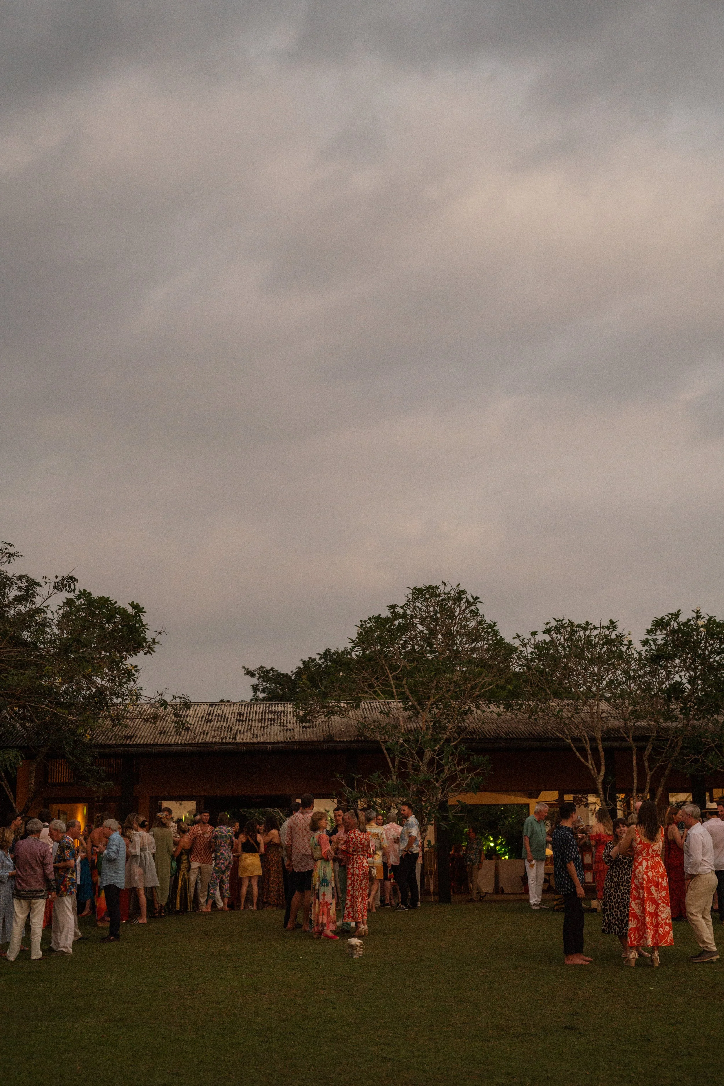 People gathered outdoors during an evening event or celebration, with some dressed in tropical or floral attire, in front of a building and trees under a cloudy sky.
