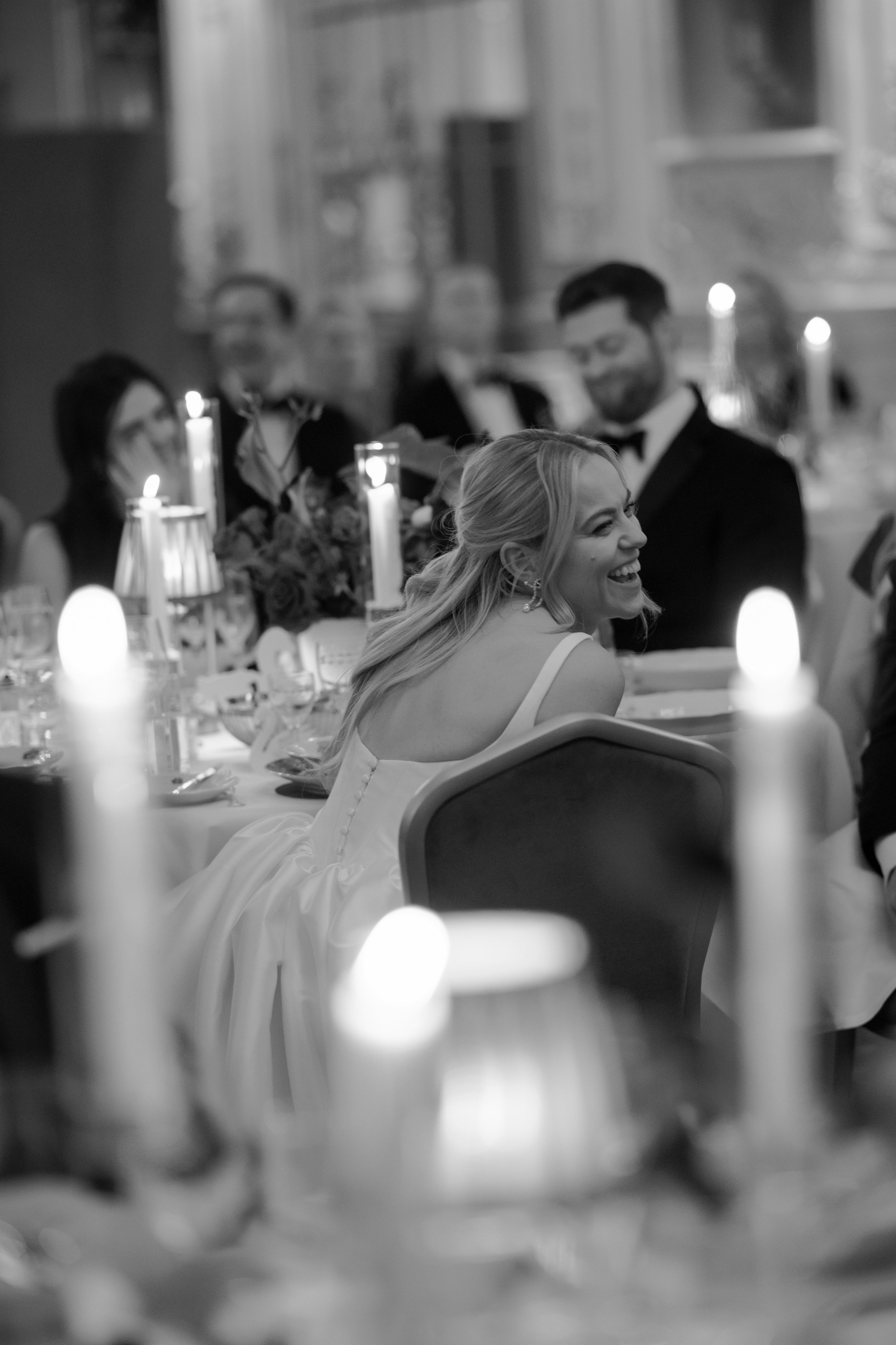 A smiling woman in a white dress sitting at a decorated table during a formal event, with people in tuxedos and elegant dresses in the background, candlelit ambiance, black and white photograph.