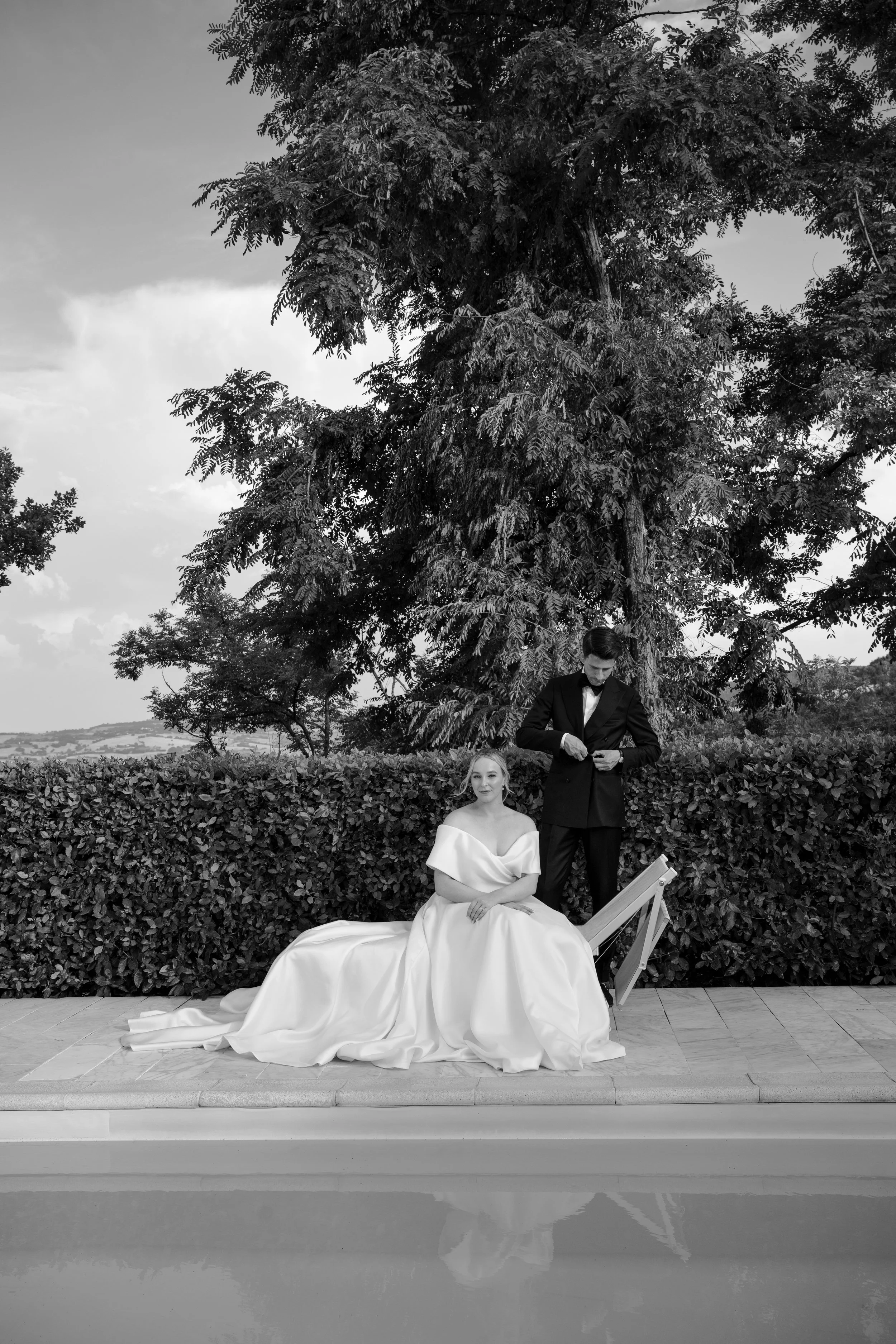 Black and white photo of a woman in a wedding dress sitting by a swimming pool, with a man in a tuxedo standing behind her, adjusting his cufflinks, outdoors with trees and a hedge in the background.