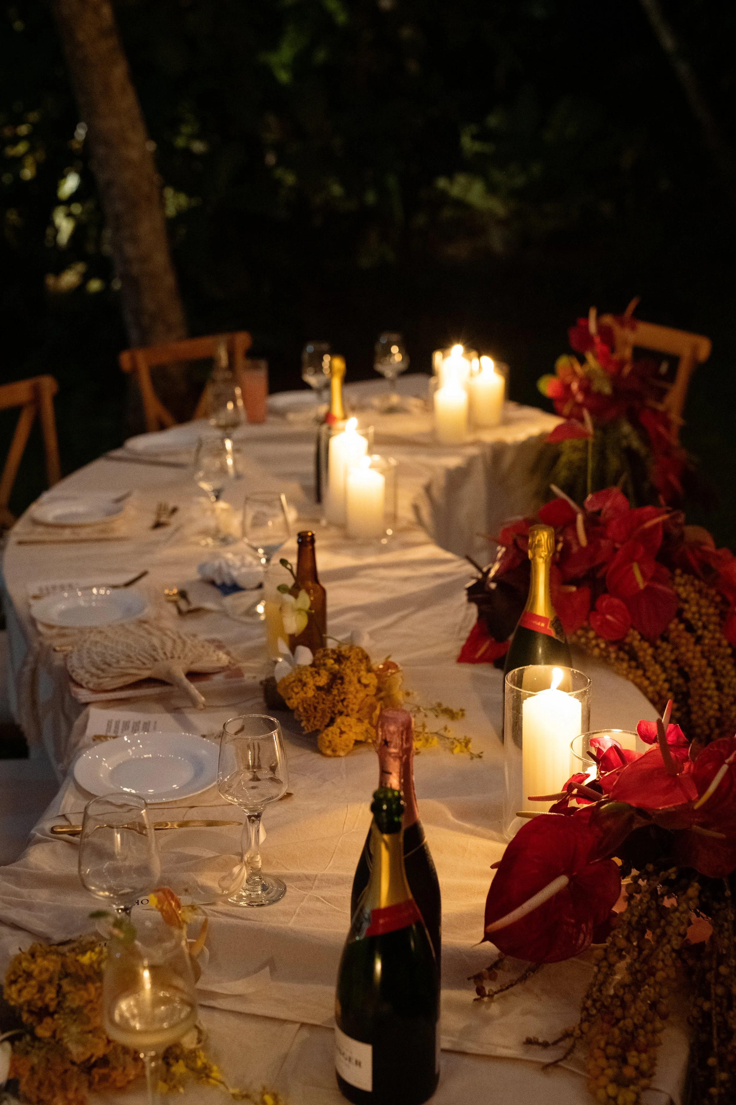 A long dining table set outdoors at night with white tablecloth, lit by candles, decorated with red flowers, and surrounded by wooden chairs.