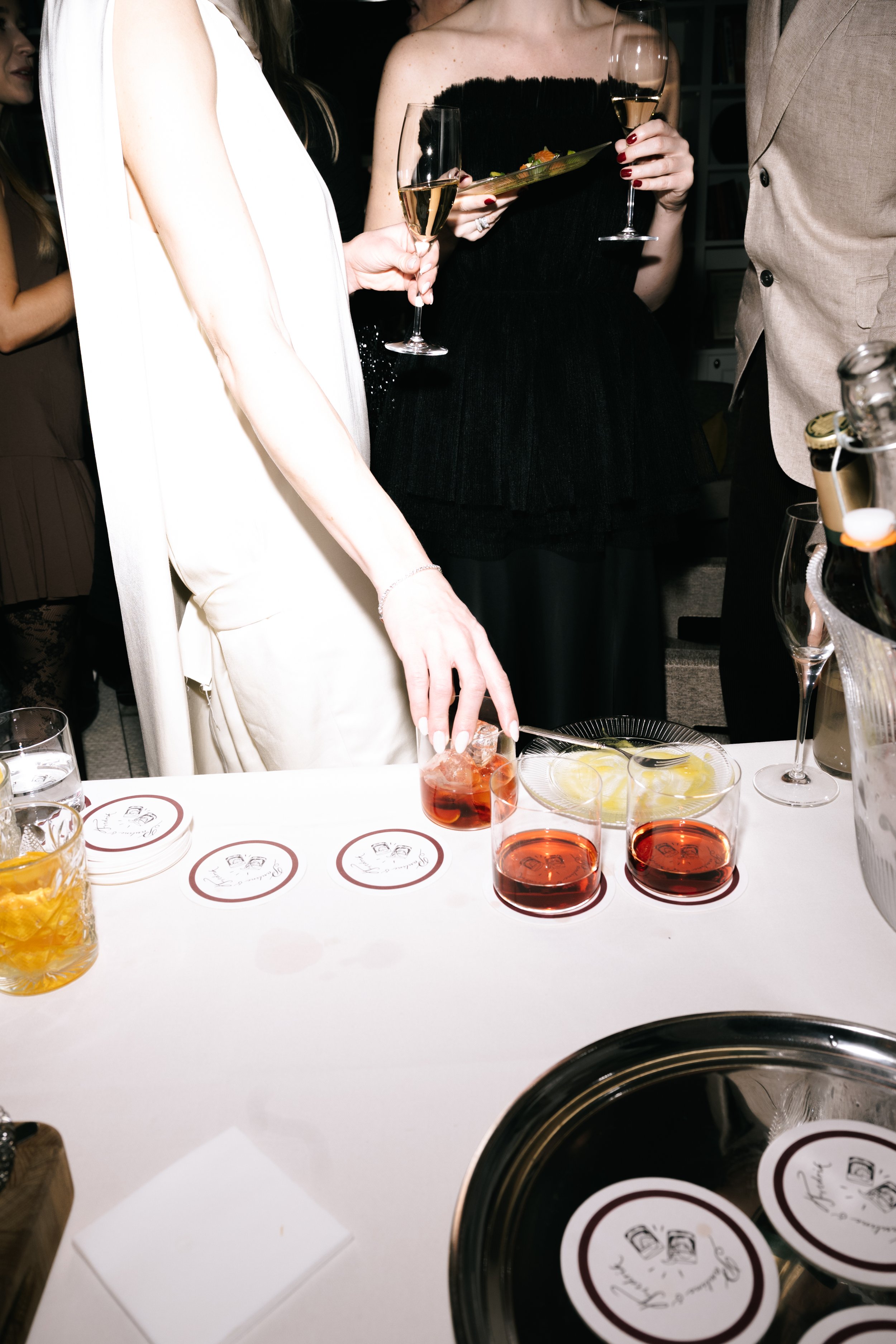 People at a social gathering preparing drinks, with wine glasses, cocktail ingredients, and coasters on the table.