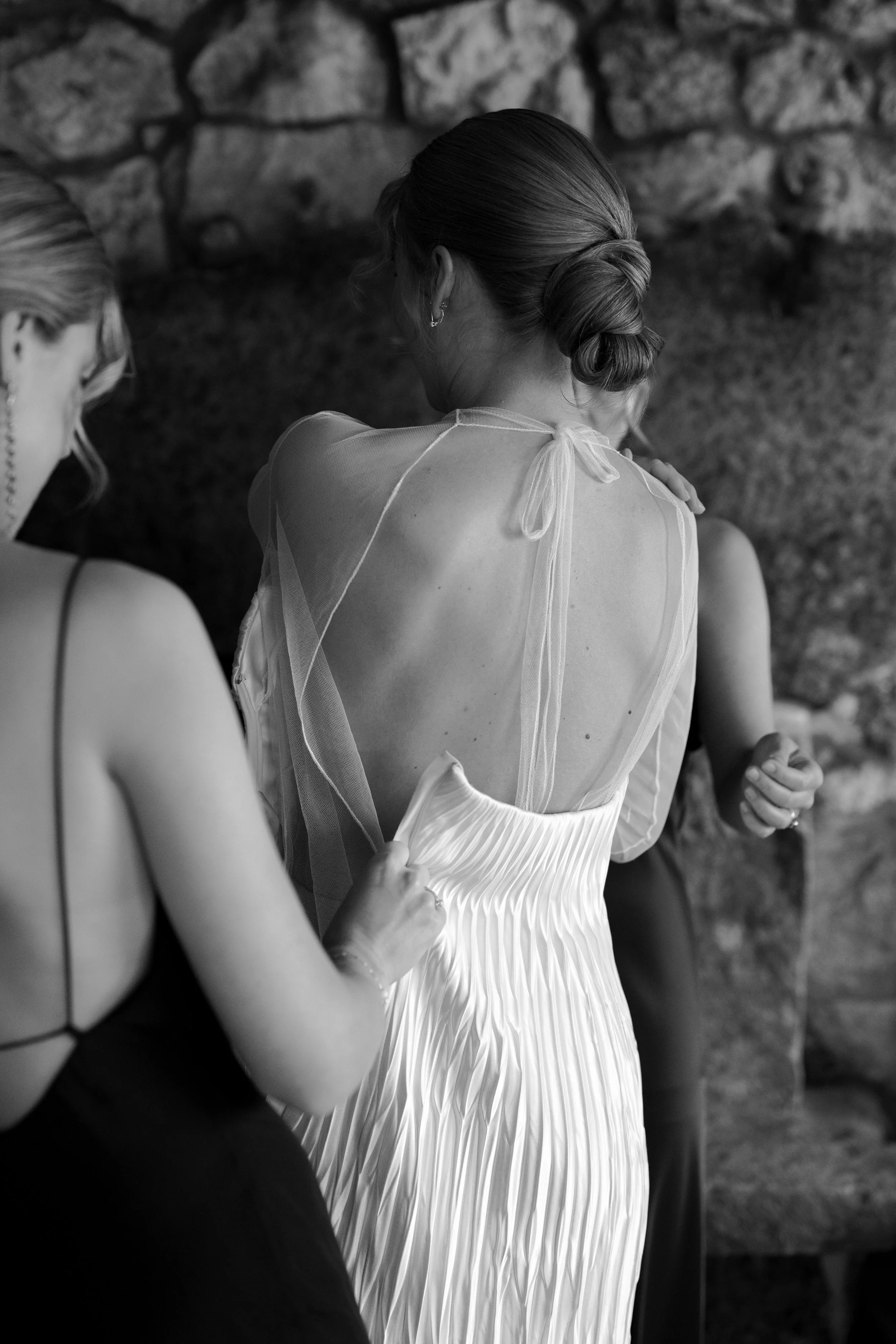 A woman in a pleated strapless dress is assisted by another woman adjusting the dress's fabric. The scene is in black and white, with a stone wall in the background.