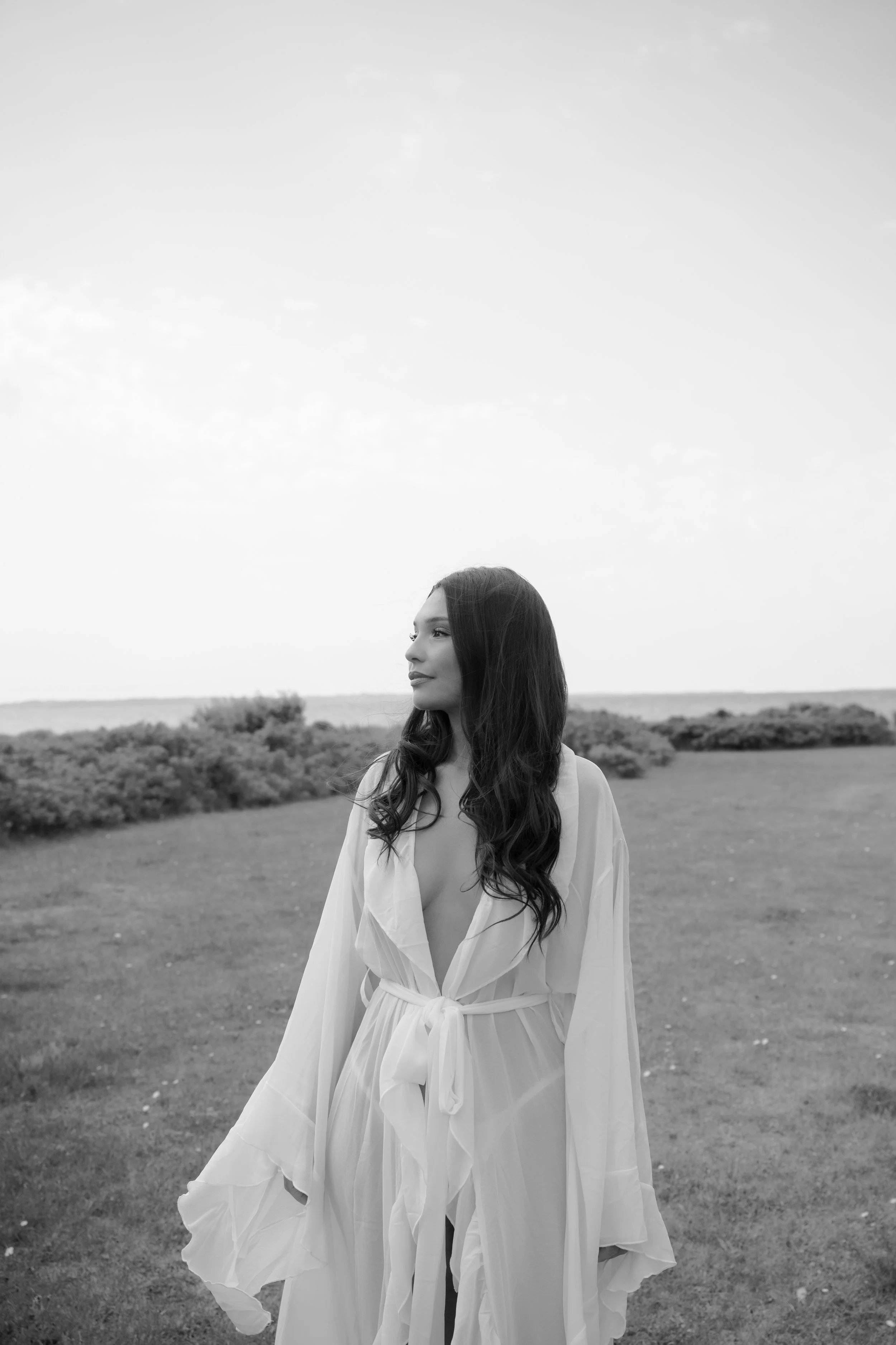 A woman with long dark hair wearing a sheer, white, long-sleeved dress with a deep neckline and a waist tie, standing outdoors on a grassy area with bushes and a body of water in the background, in black and white.