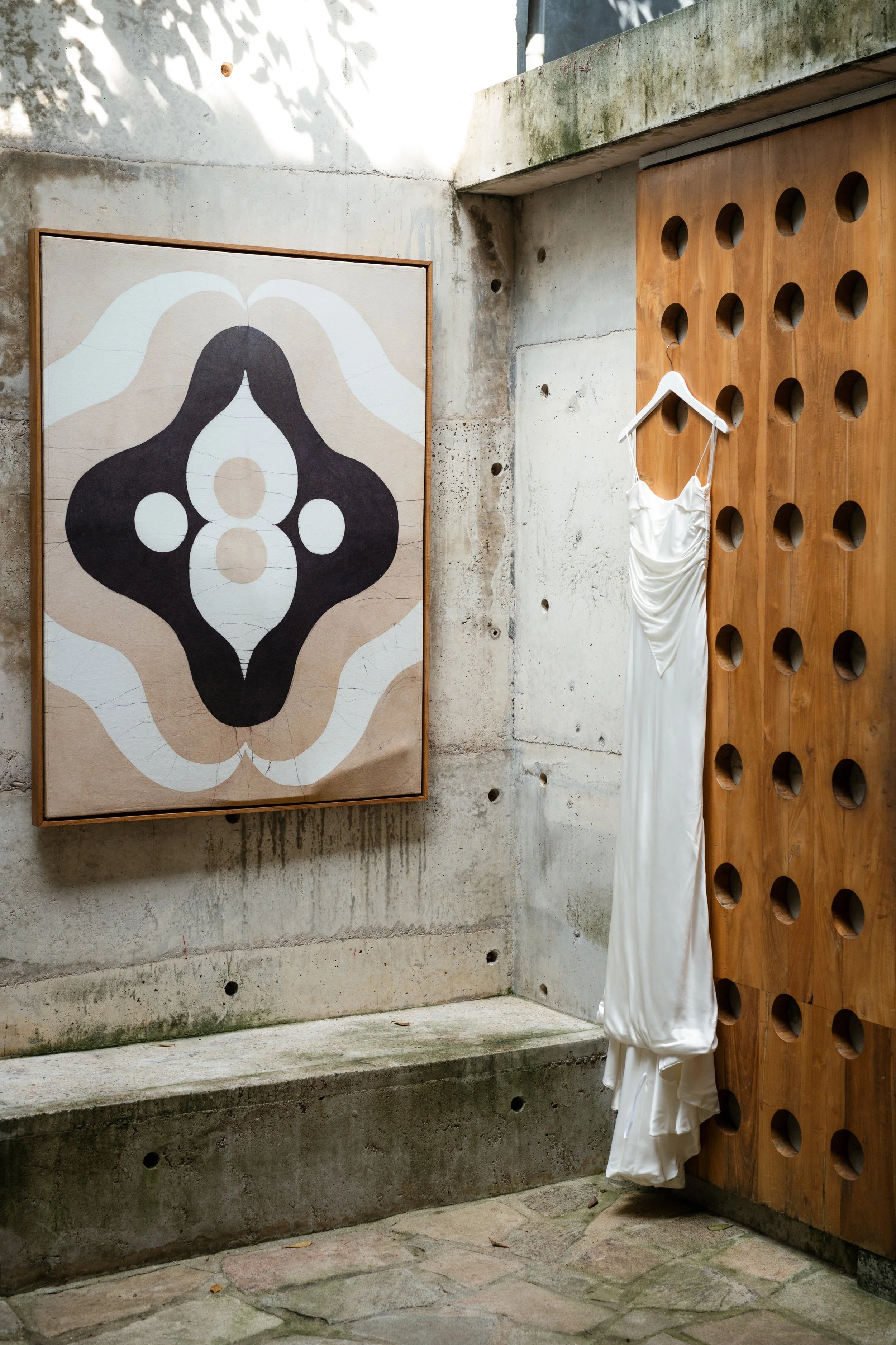 A white dress hanging on a wooden wall with circular holes, next to an abstract painting on a concrete wall.