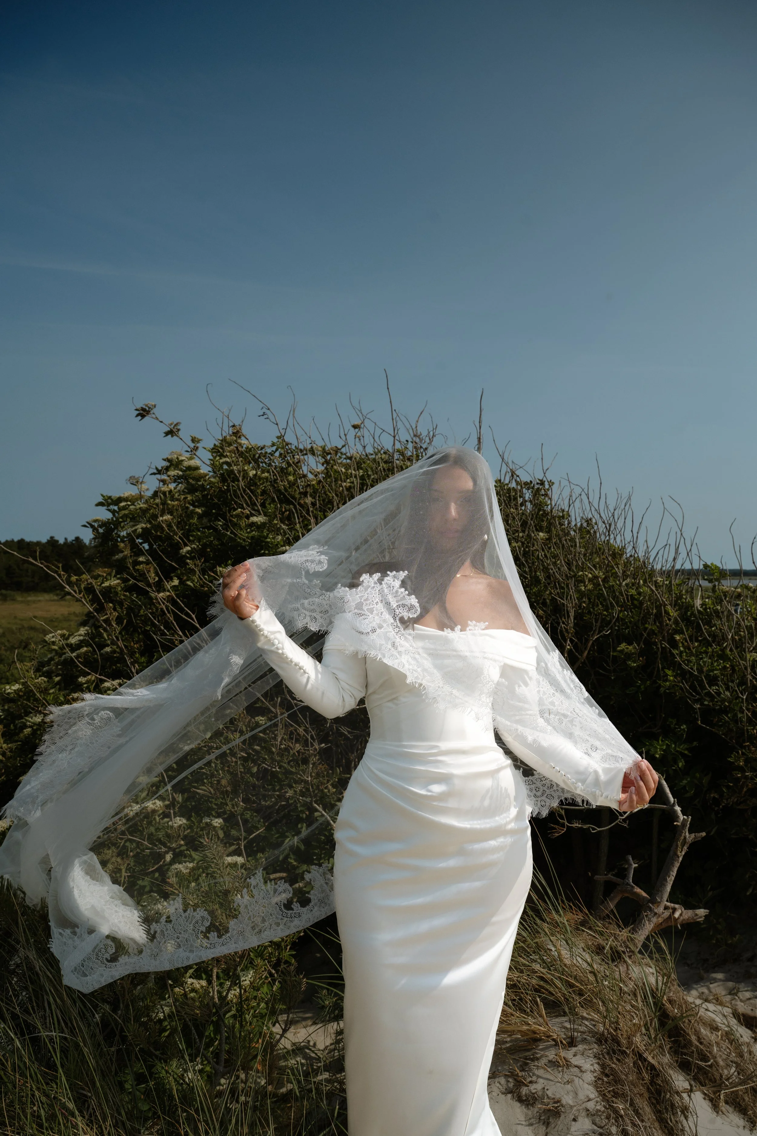 A woman in a white dress with long sleeves standing outdoors with a veil over her face, holding the veil with one hand. She is positioned in front of green foliage and a clear blue sky.