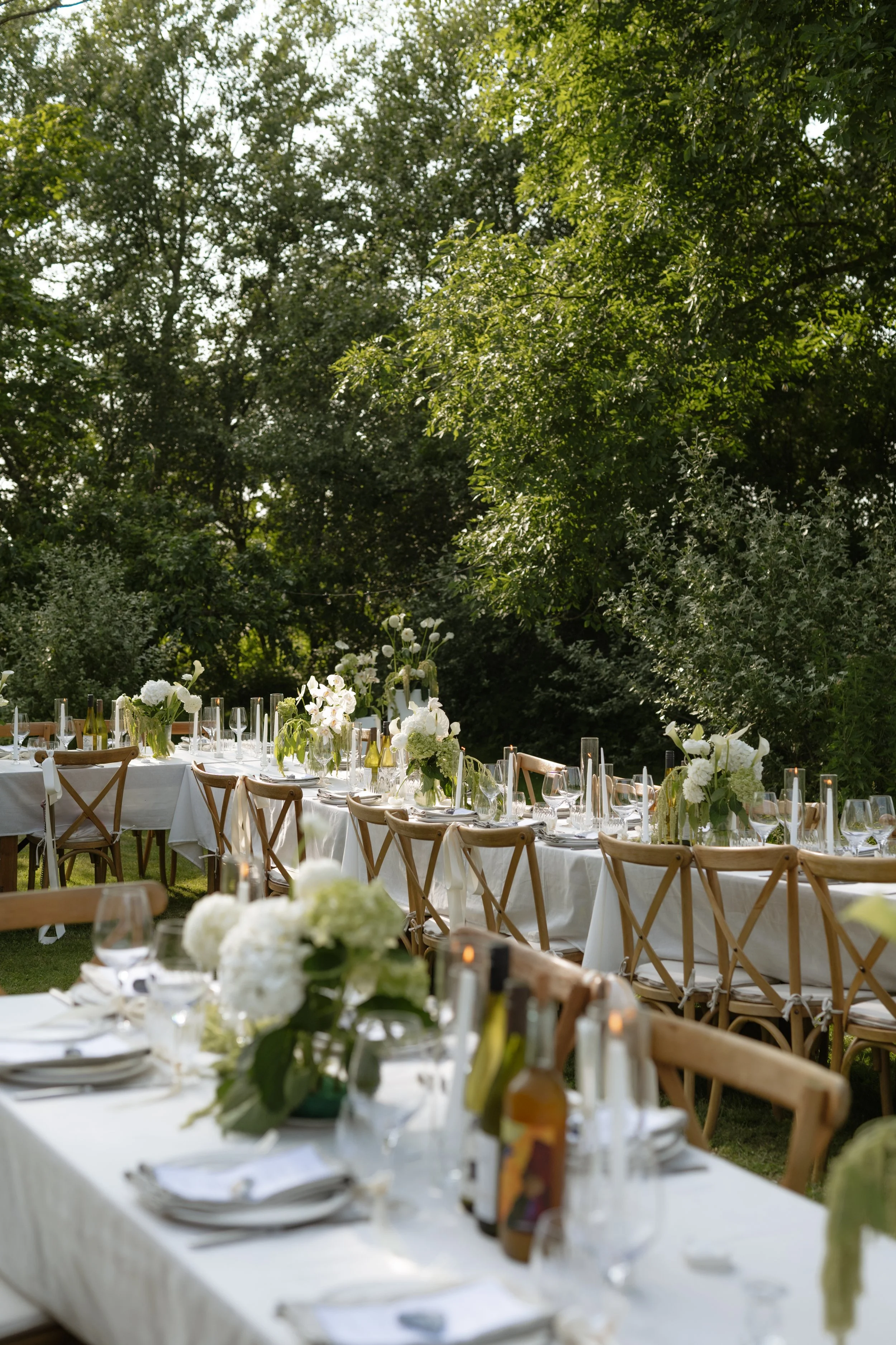 Outdoor dining table set up for an event with white tablecloths, floral centerpieces, and wine glasses, surrounded by chairs with crisscross backs, in a lush green garden.