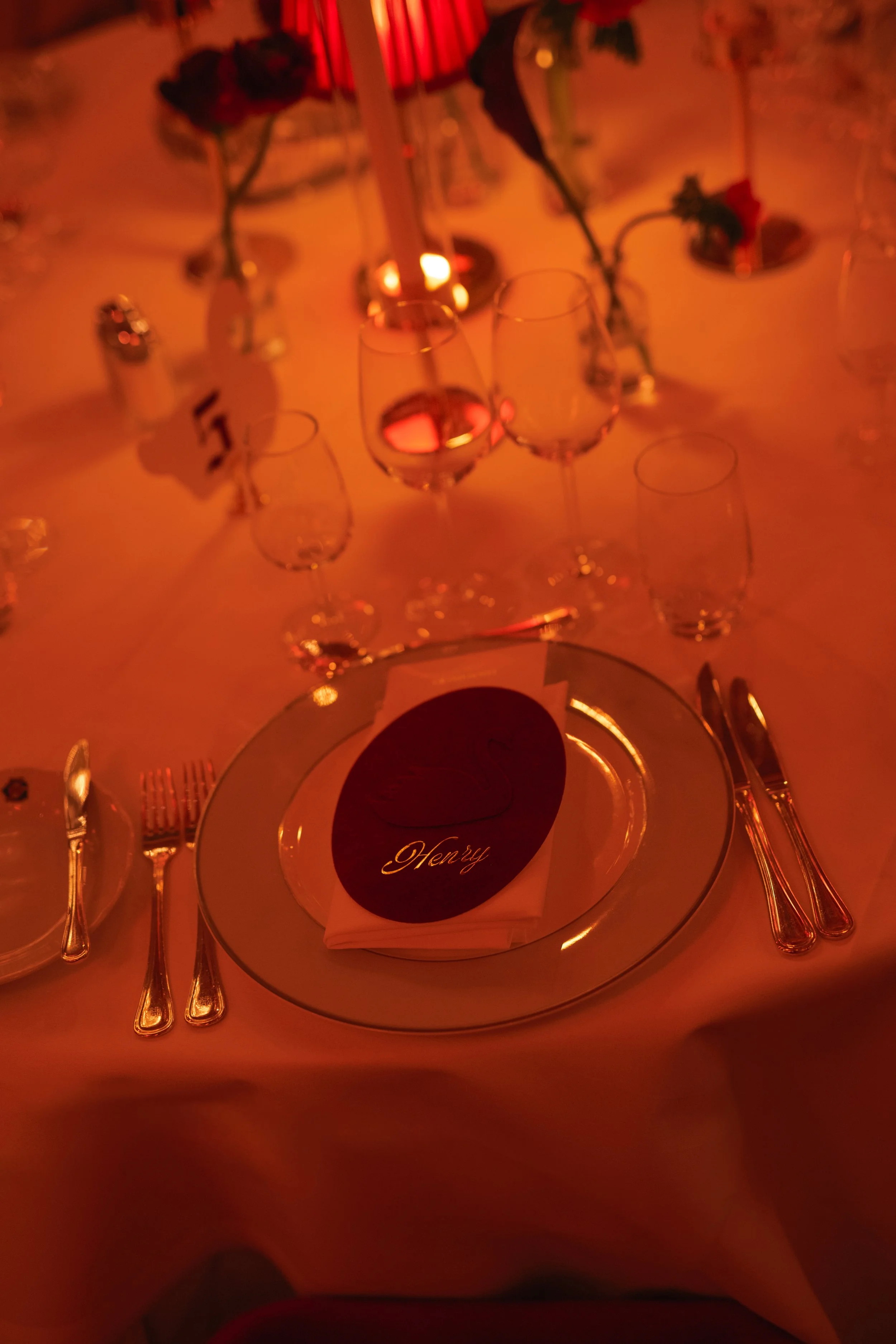 A formal dining table set with wine glasses, silverware, and a napkin with a place card that says 'Henry,' under warm orange lighting with floral centerpieces.