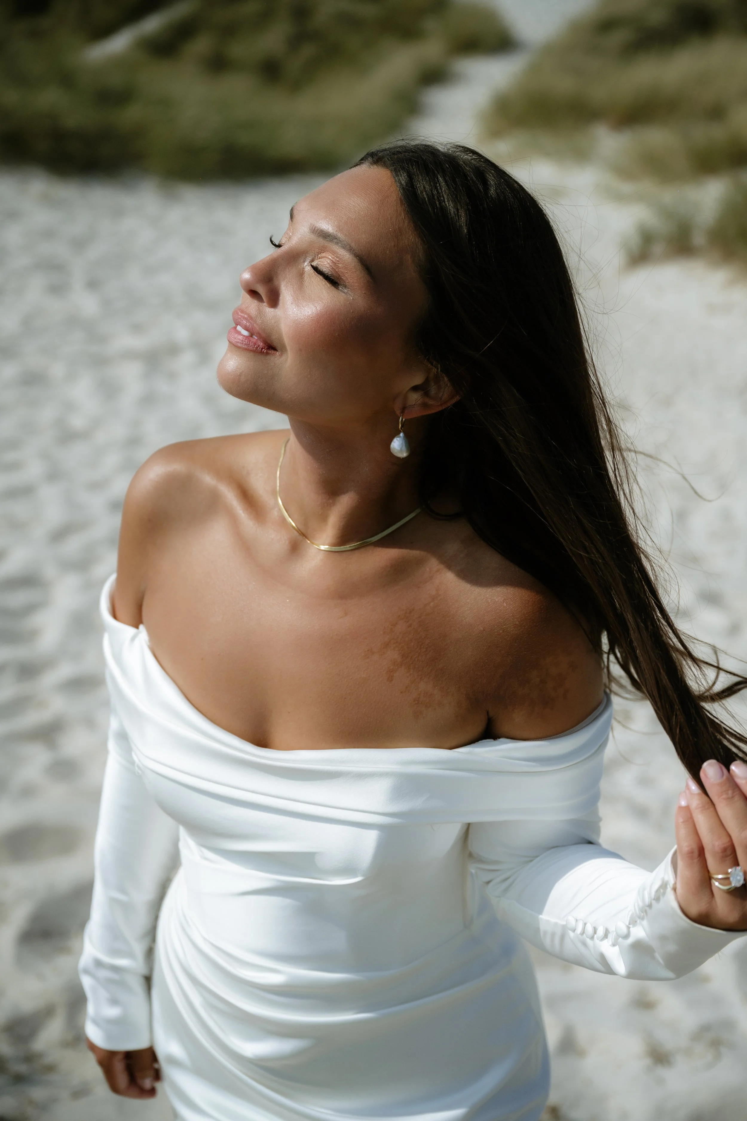 Woman with long dark hair in a white off-shoulder dress standing on a beach with her eyes closed and face uplifted to the sun.