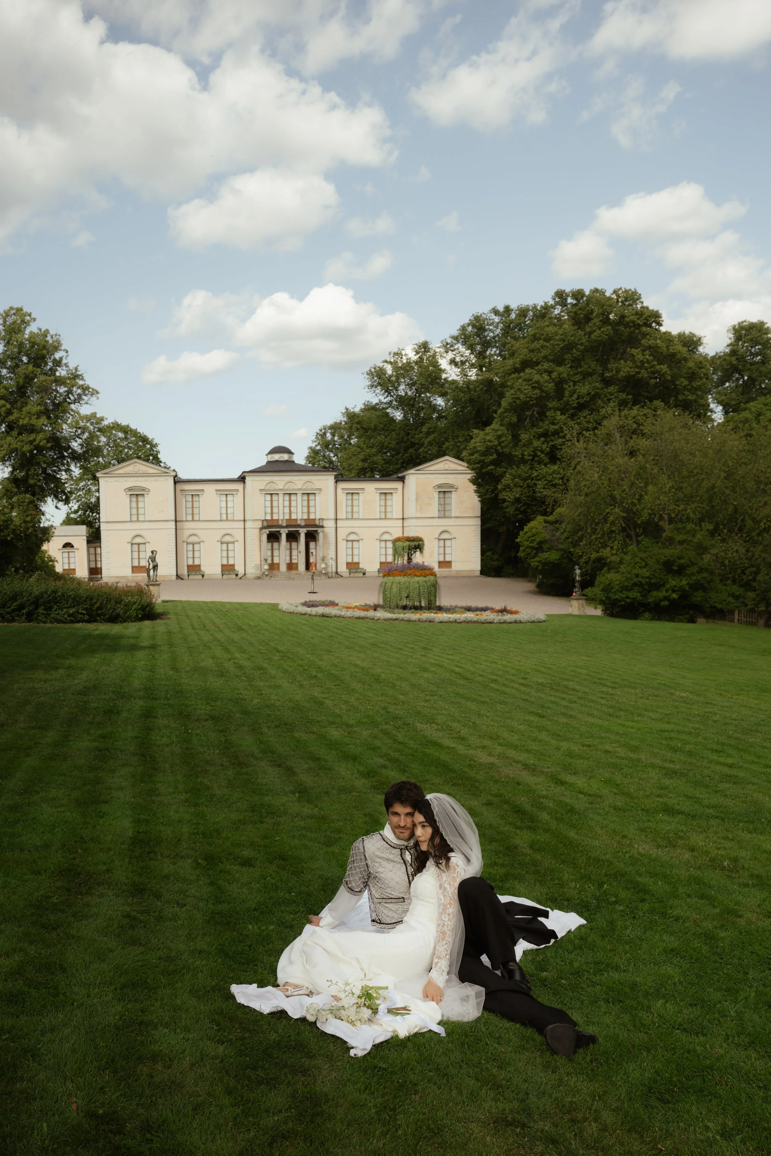A bride and groom sitting on the grass with a grand estate in the background.