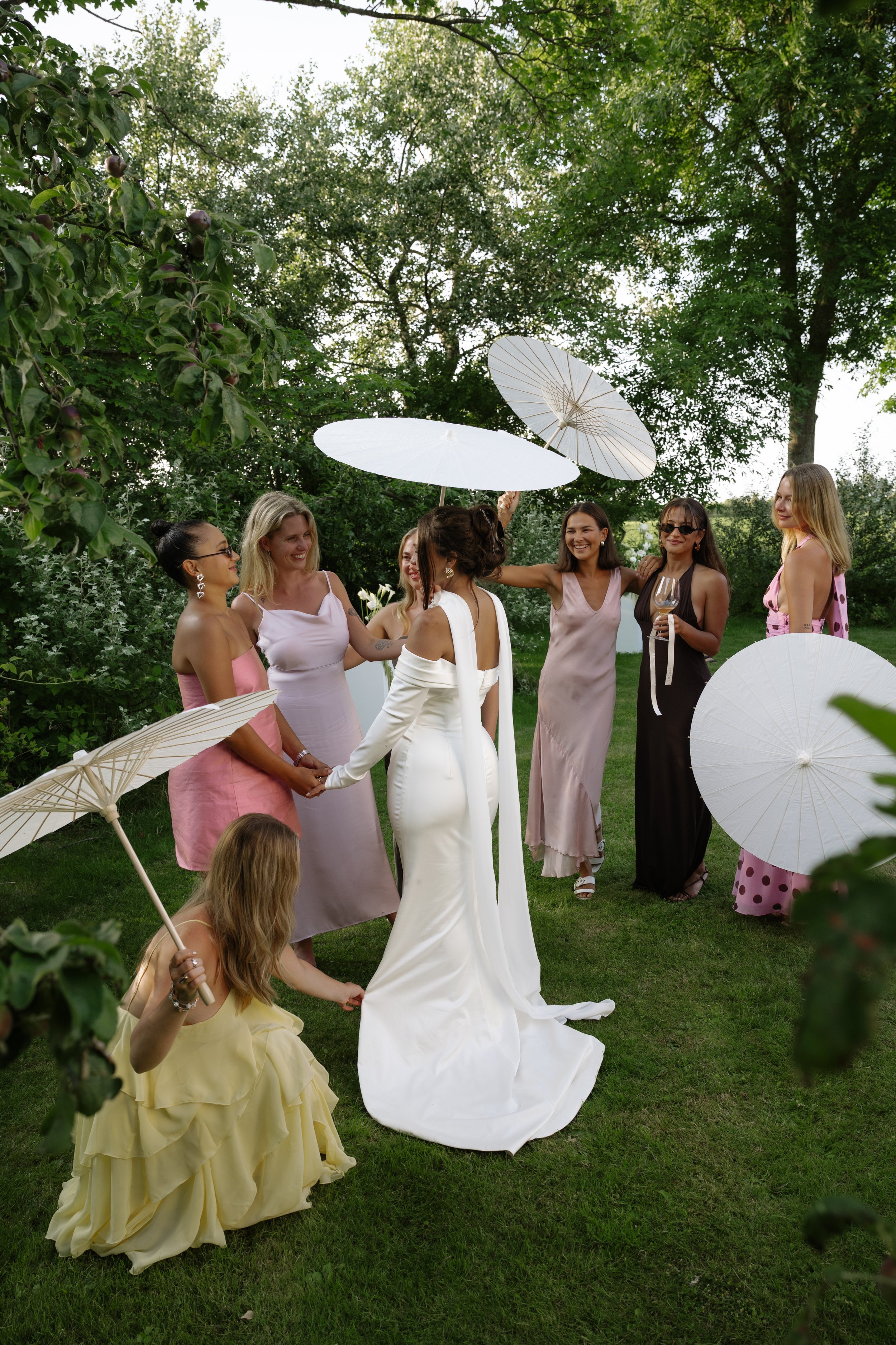 A group of women at a wedding outdoors, holding umbrellas, with one woman in a white wedding dress and others in colorful dresses, standing on green grass surrounded by trees.