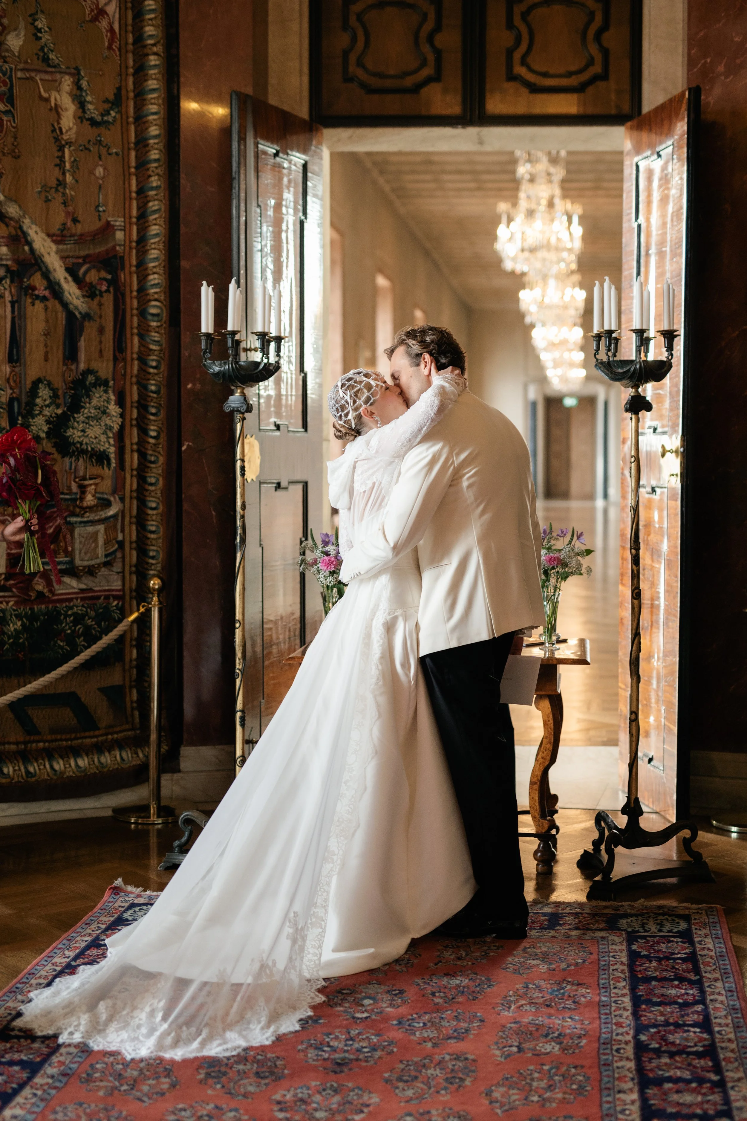 A bride and groom share a kiss in a grand, elegant hallway with chandeliers and vintage decor, with the bride in a white gown and the groom in a white tuxedo jacket.