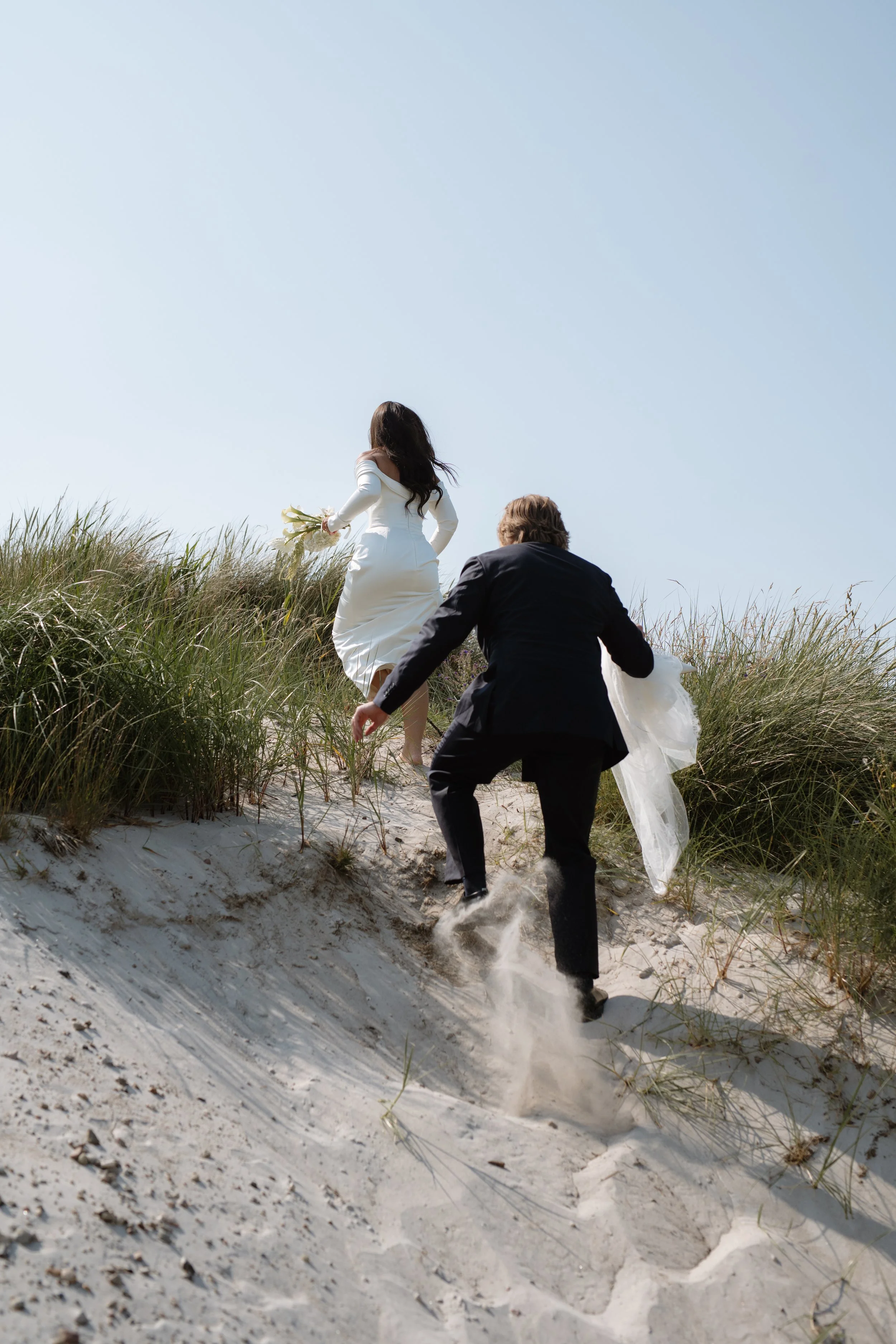 A couple in formal wedding attire running up a sandy dune on a beach with tall grass, under a clear blue sky.