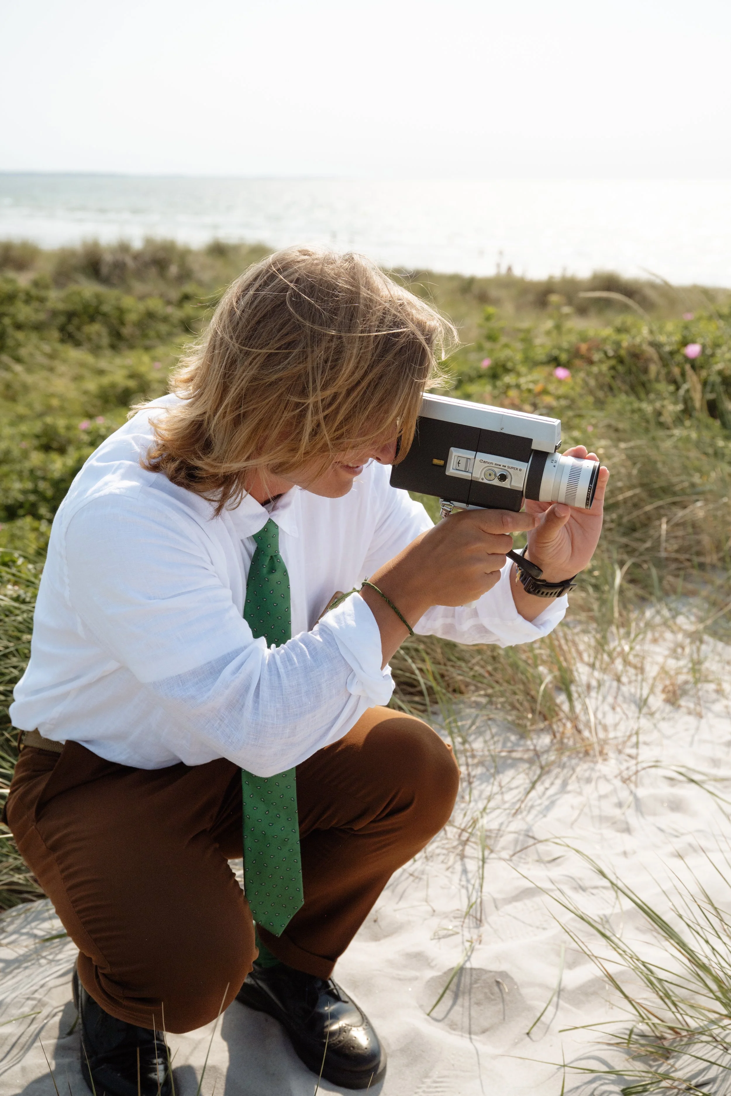 A man with shoulder-length blonde hair in a white shirt and green polka-dot tie crouches on sandy beach grass, looking through a vintage black and silver camera.