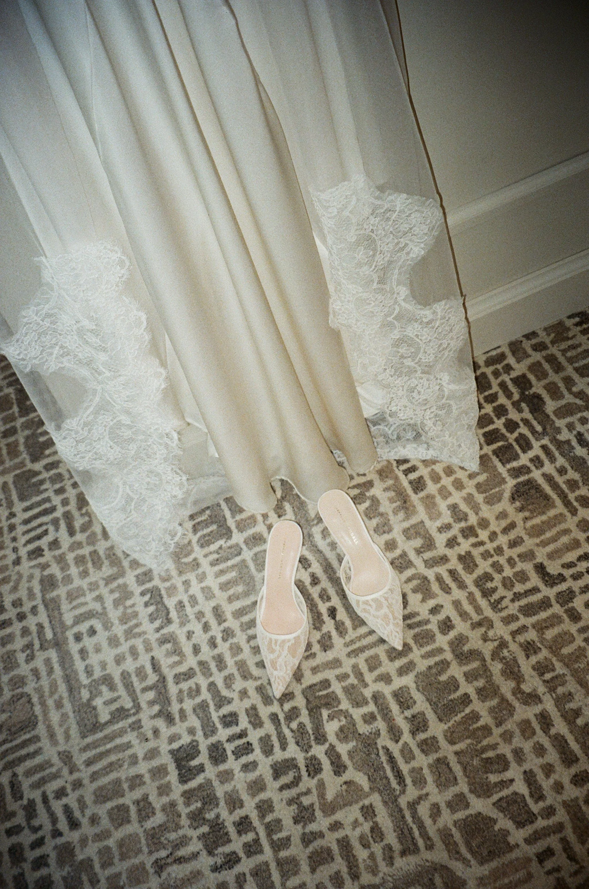 A pair of cream high-heeled shoes with lace pattern, placed on a patterned carpet next to a cream-colored dress with lace details.