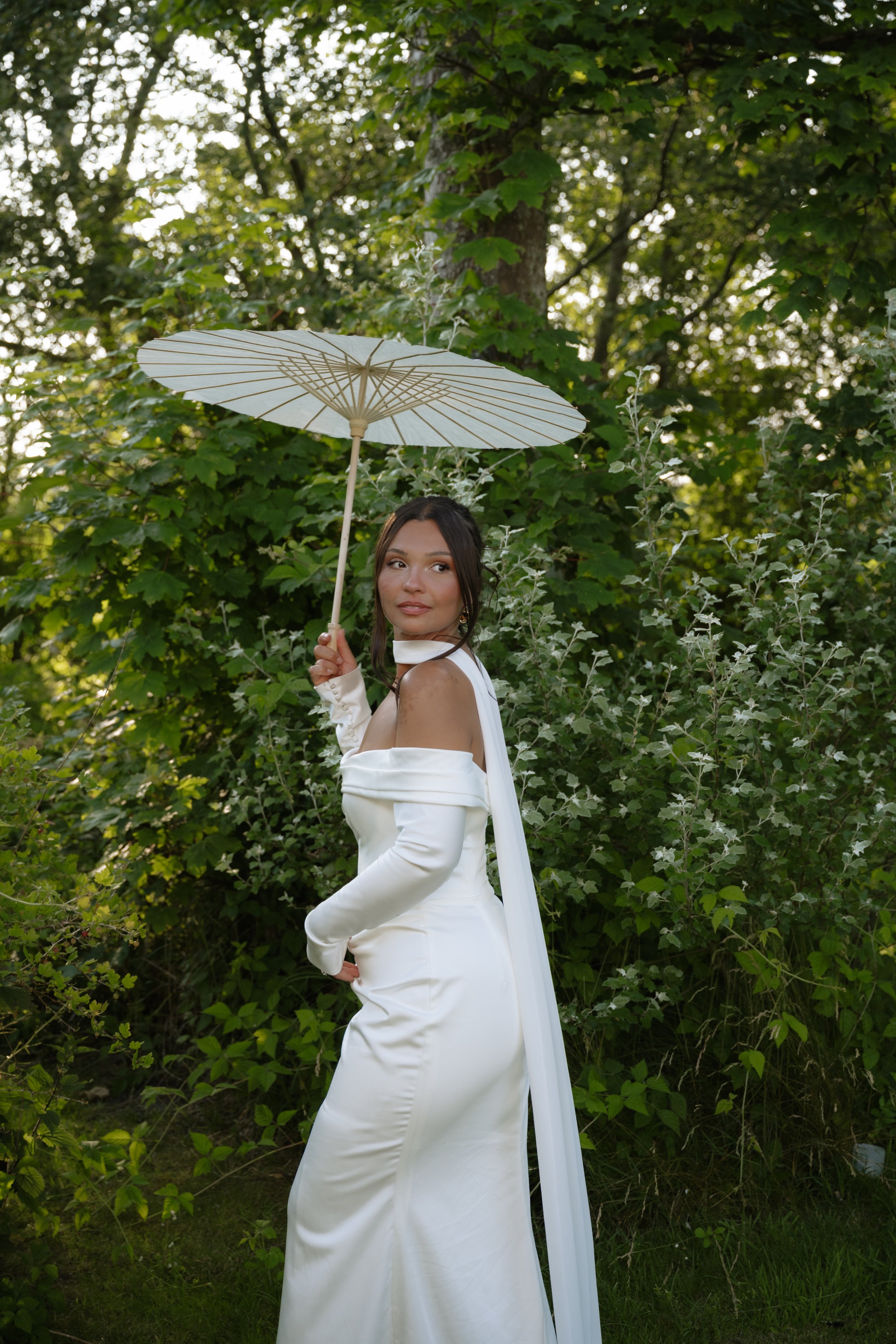 A woman wearing a white dress holding a white parasol in a lush green outdoor setting.