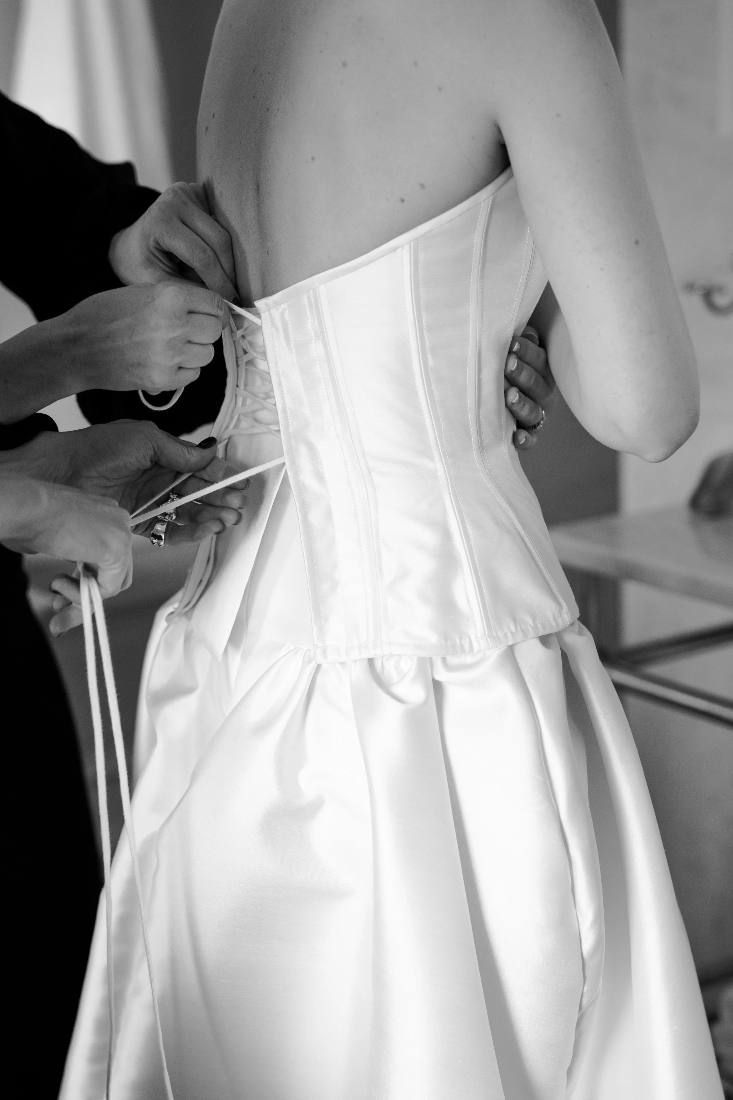 A bride is wearing a satin wedding dress while having a corset back laced up by someone.