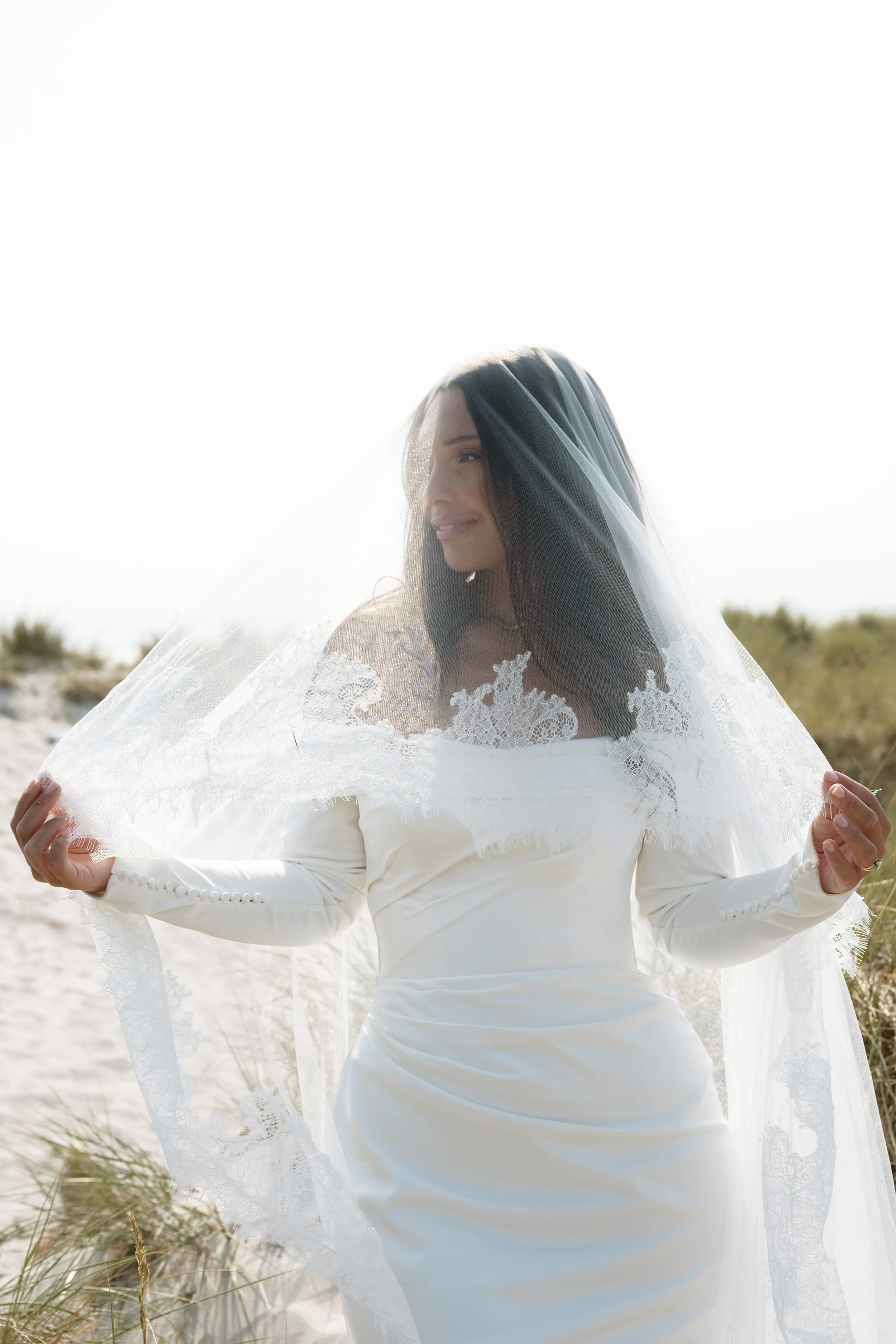 A woman in a white wedding gown with a veil standing outdoors on a sandy beach with grassy dunes, smiling gently with the sunlight backlighting her.