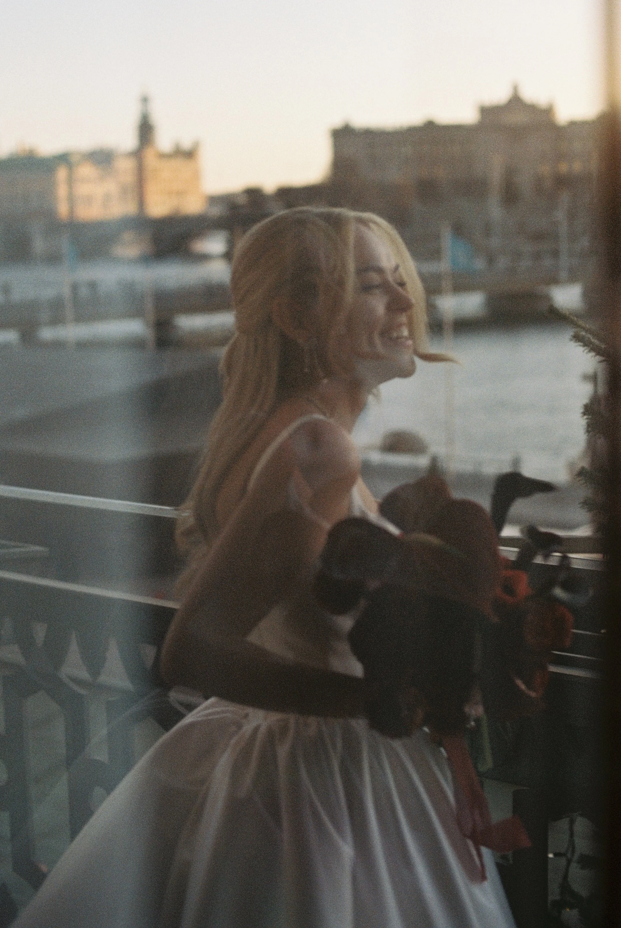 A smiling woman with blonde hair wearing a white dress, holding a bouquet of flowers, seen through a window with a city and water in the background.