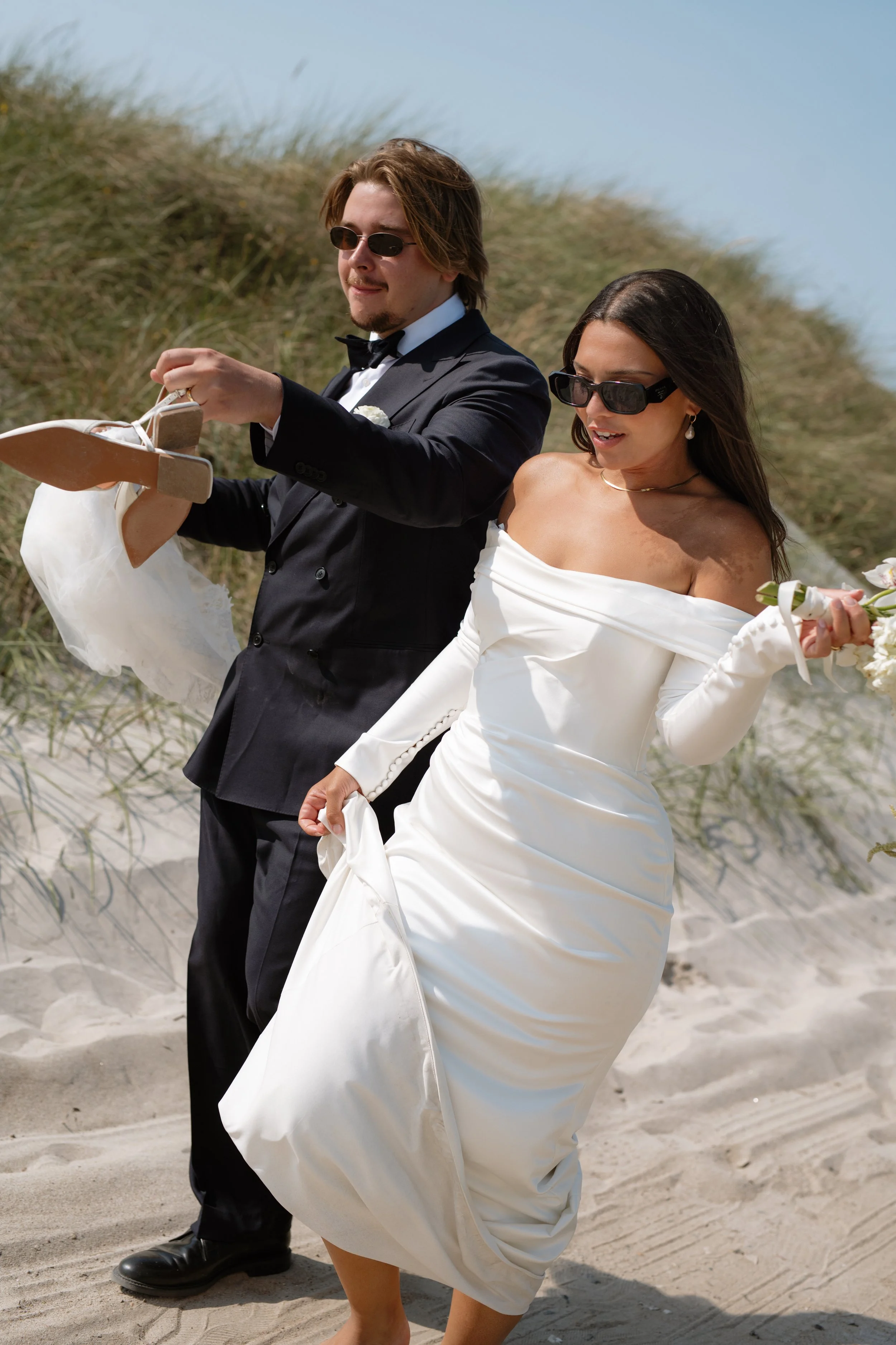 A bride and groom walking barefoot on the beach, both wearing sunglasses; the groom is in a black tuxedo and the bride is in a white off-shoulder wedding dress, holding flowers.
