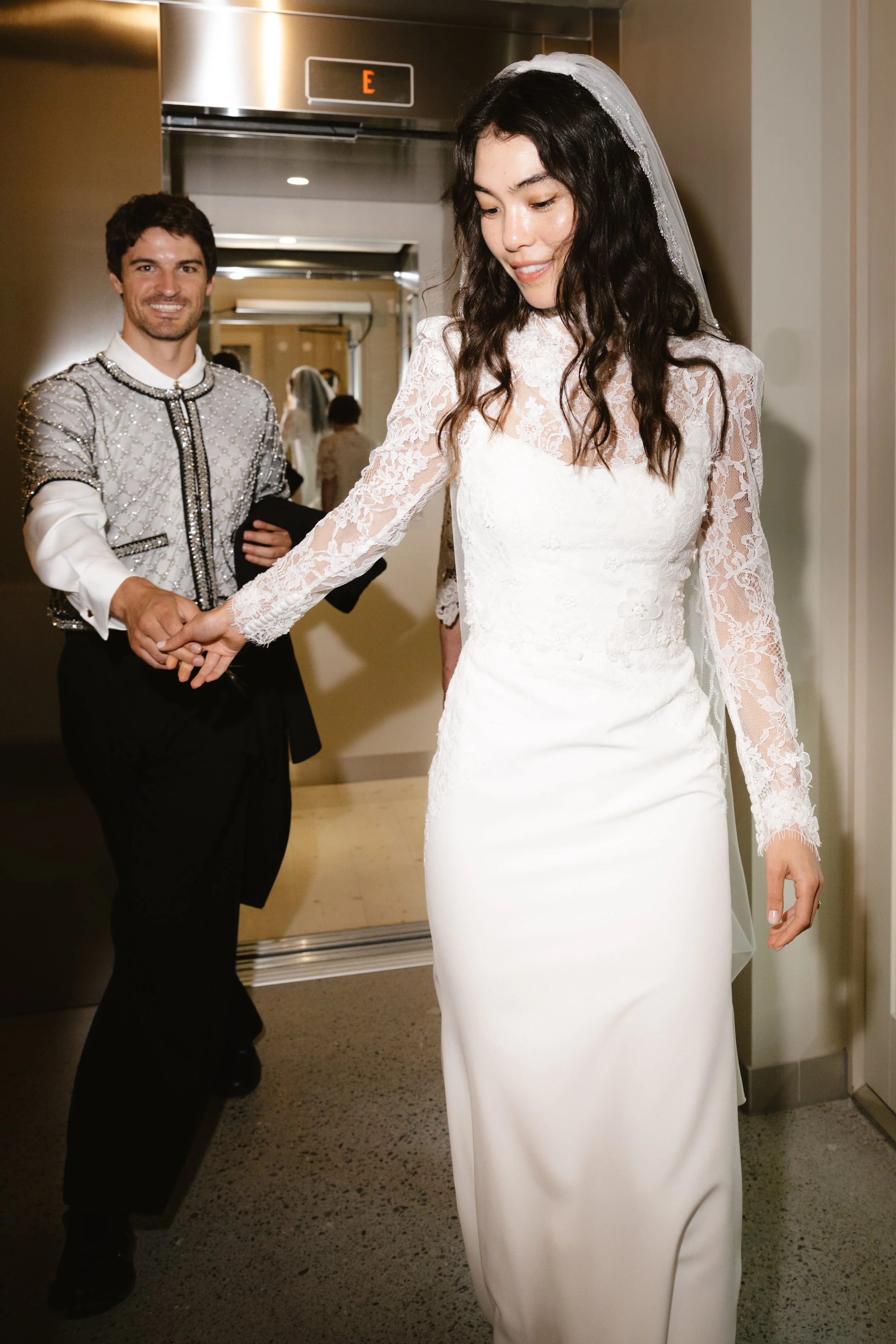 A bride in a white lace wedding dress with a veil is holding hands with a groom in a silver and black tuxedo as they exit an elevator.
