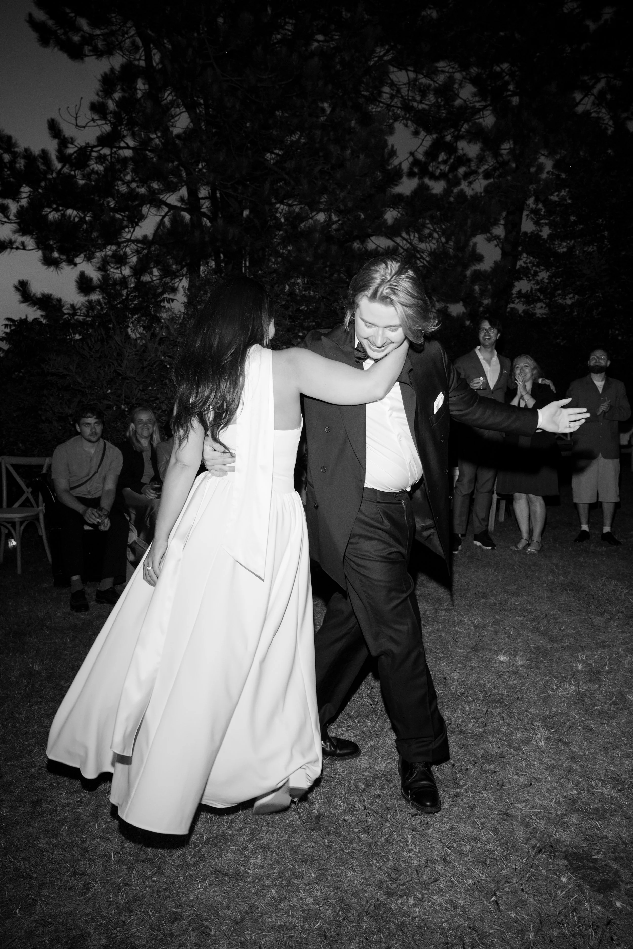 A black and white photo of a wedding reception. A bride in a white gown and a groom in a tuxedo are dancing together, smiling, with the bride's arm around the groom's neck. In the background, guests are watching and smiling, standing outdoors near tr