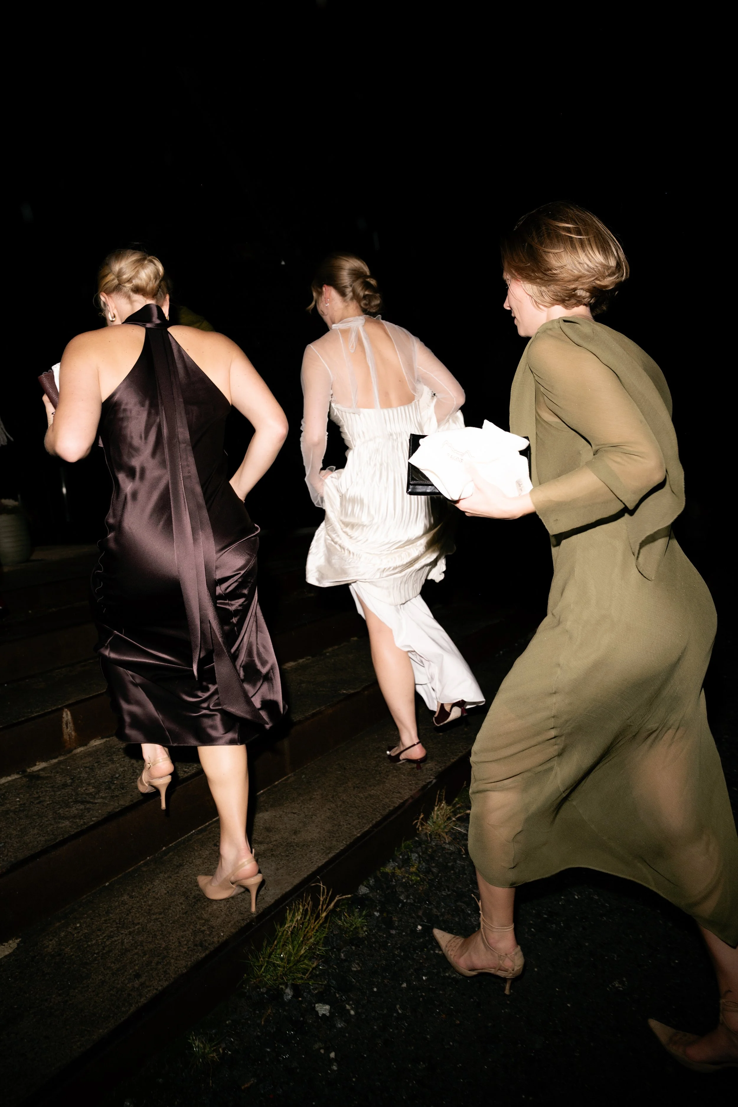 Three women walking up a set of outdoor stairs at night, dressed in elegant evening attire, one holding a white cloth or dress and a small black purse.
