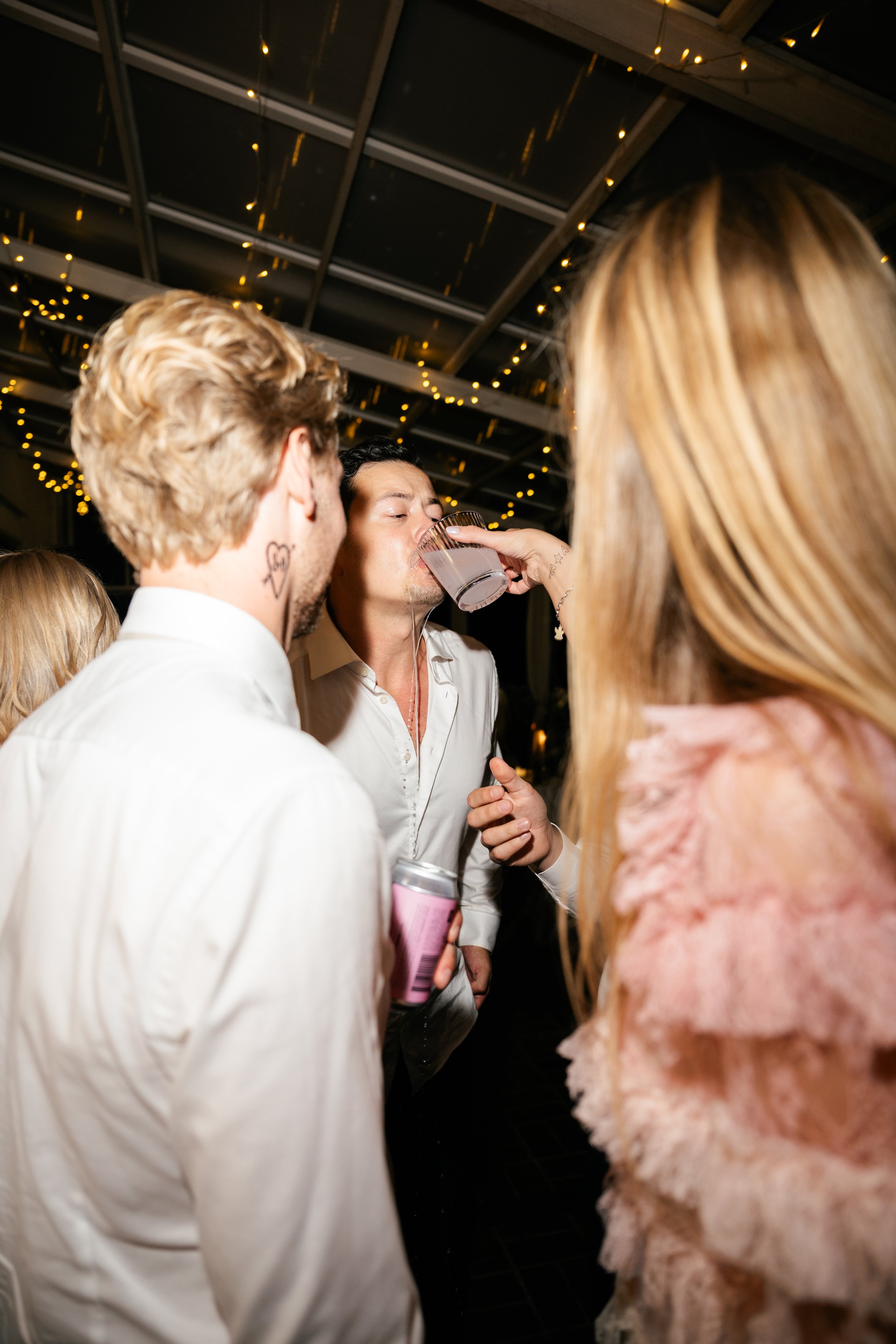 People at a party, with a man drinking from a glass held by a woman, surrounded by others, with string lights overhead.