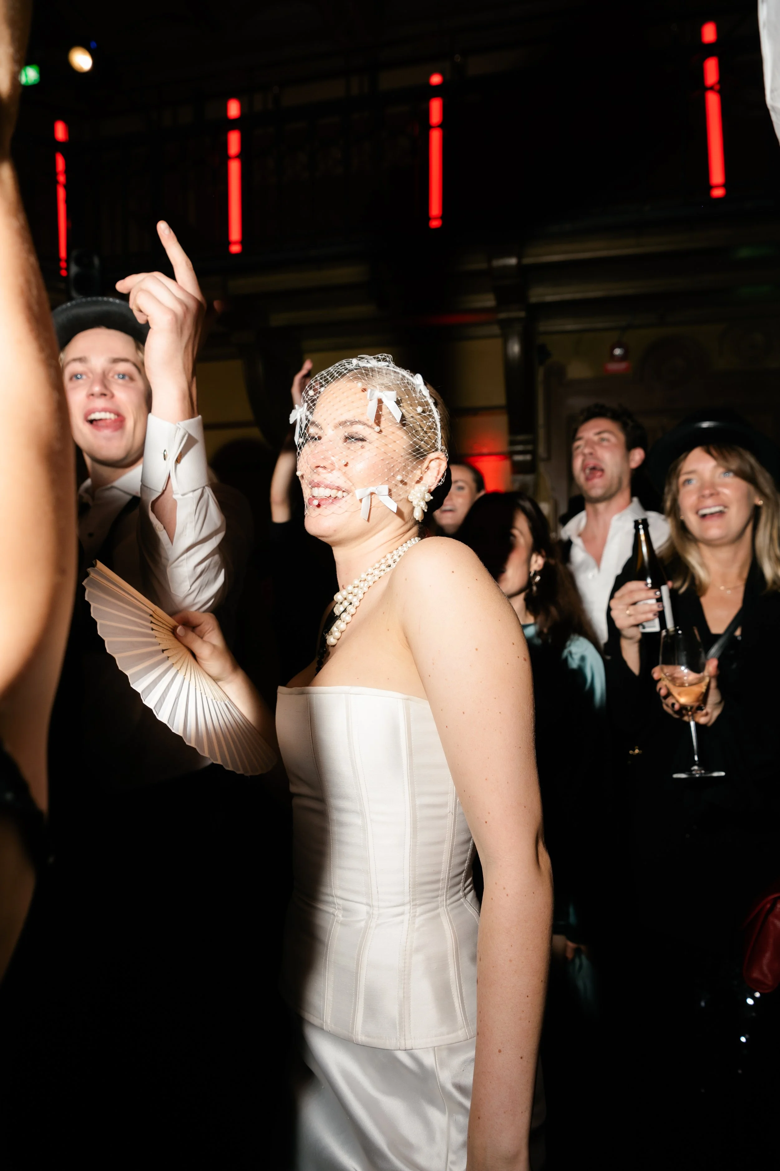 A smiling woman in a white strapless dress and pearl necklace, wearing a birdcage veil, at a lively party surrounded by guests.