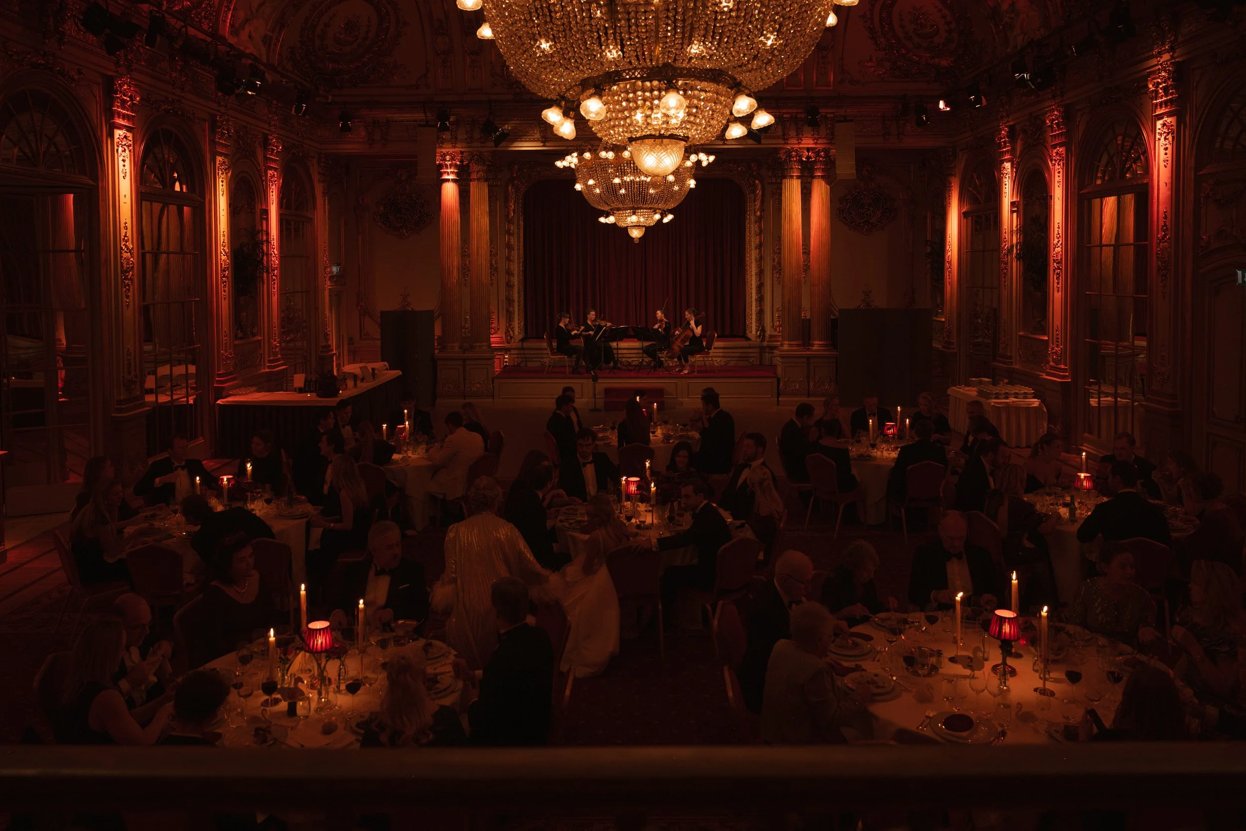 An elegant banquet hall with ornate wood paneling and large chandeliers, filled with round tables set for dinner, and a live classical music performance on a stage at the front of the room.
