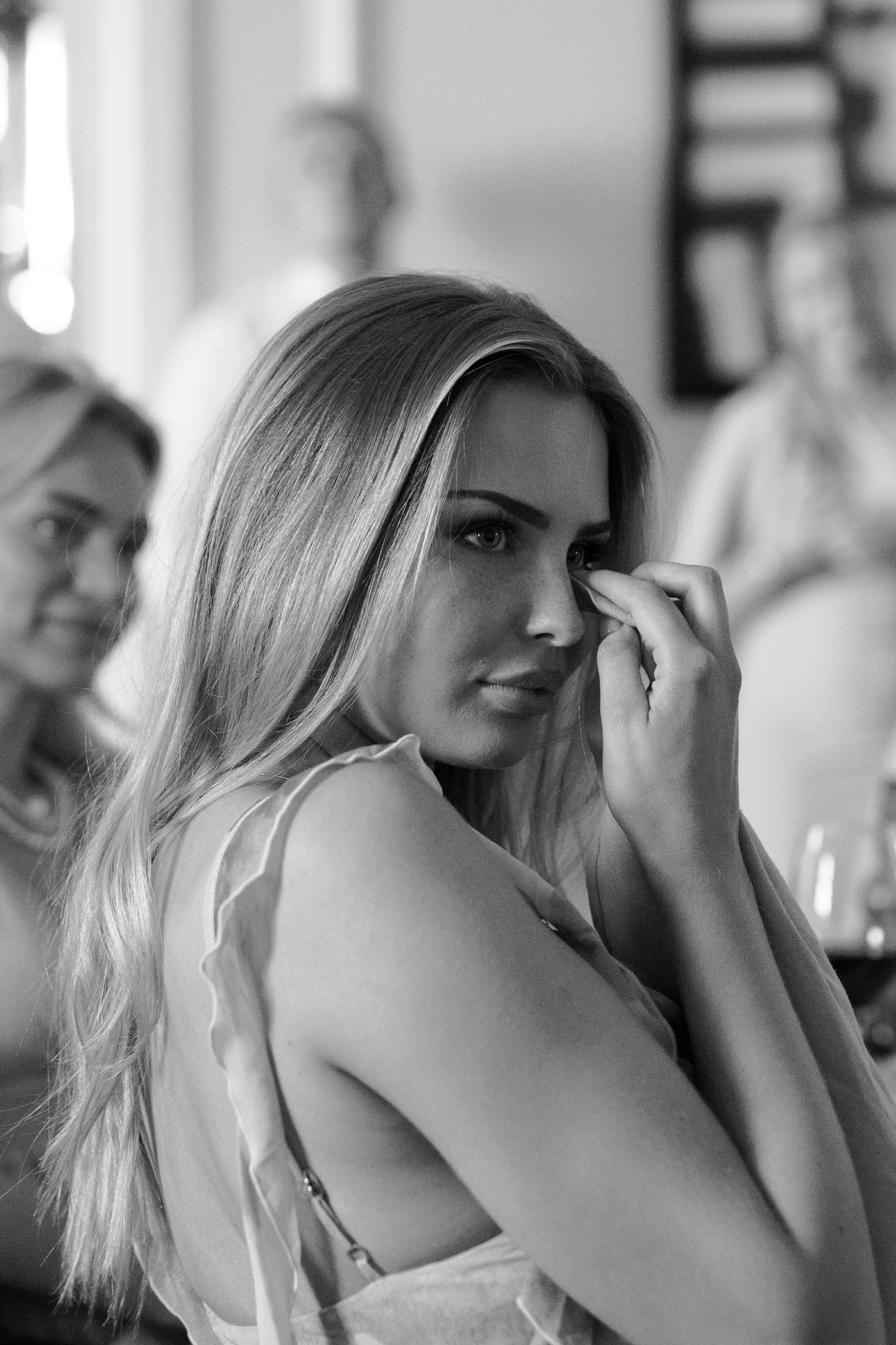 A woman with long hair touching her eye, seated in a social setting with other women in the background.