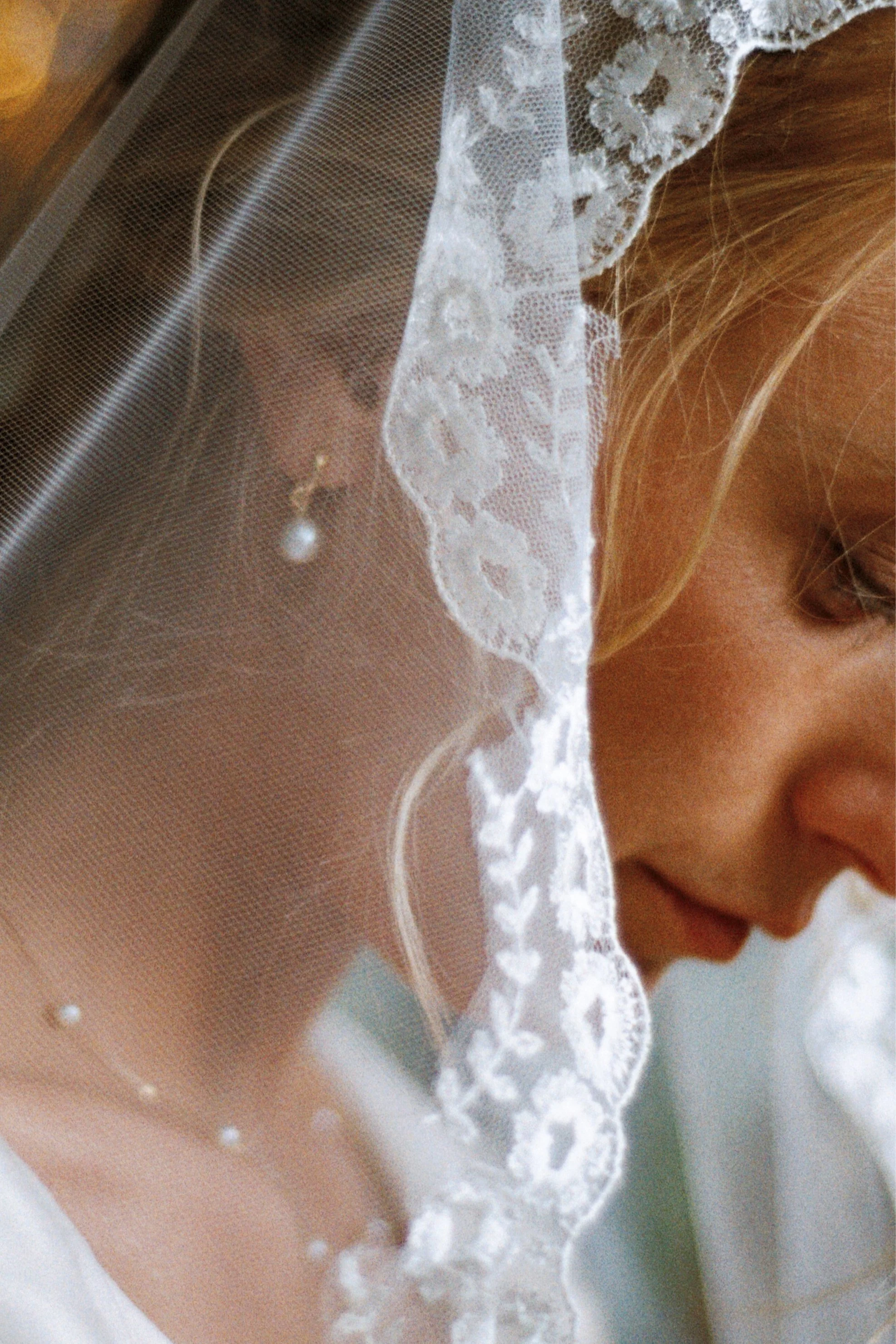 Close-up of a bride with a lace veil, wearing a pearl earring, with her face slightly turned down.