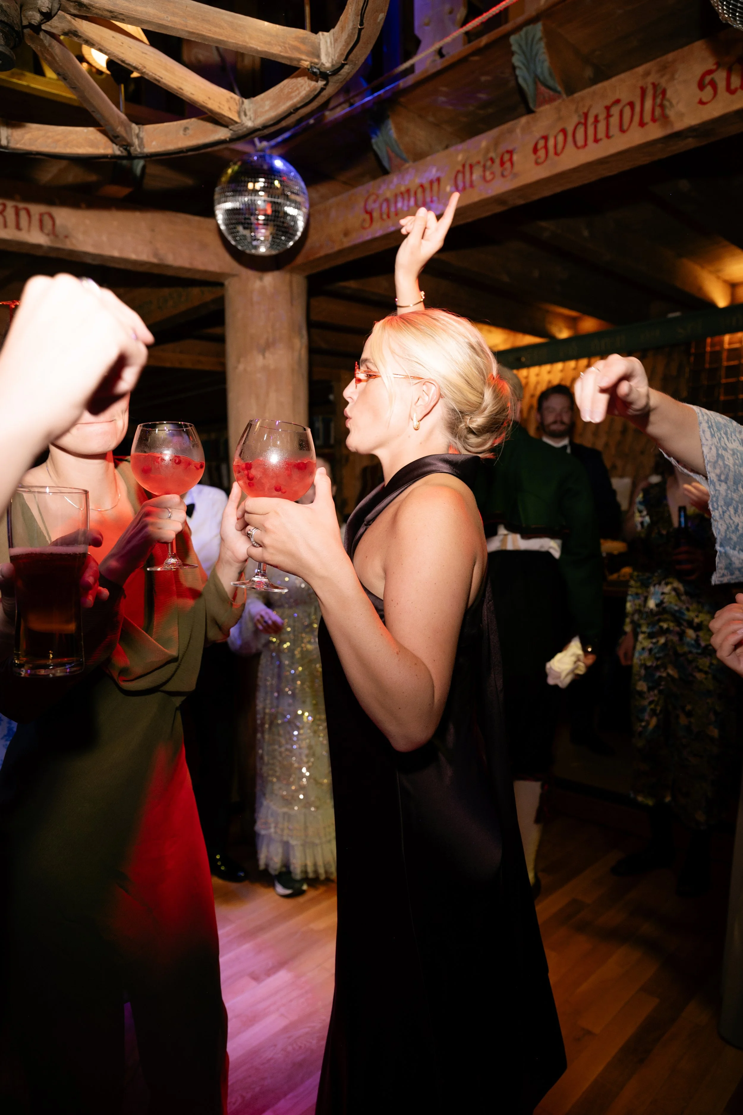 A woman with blonde hair and glasses at a party, holding a glass of pink cocktail with red berries, surrounded by other guests, in a rustic venue with wooden beams and a disco ball.