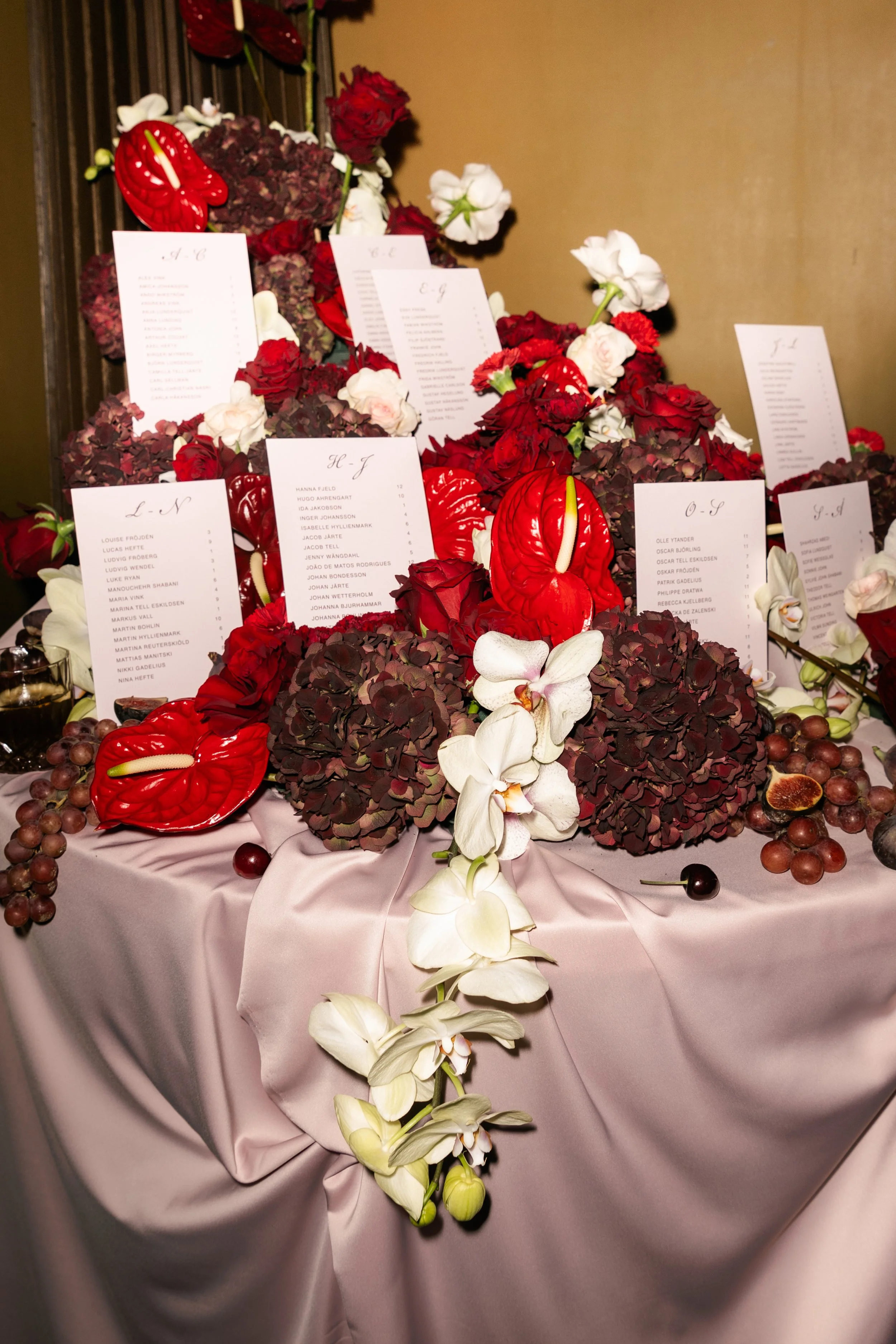 A floral wedding seating chart arrangement with red, white, and purple flowers, including orchids and hydrangeas, on a pink satin tablecloth, with cards displaying guest names and table assignments.