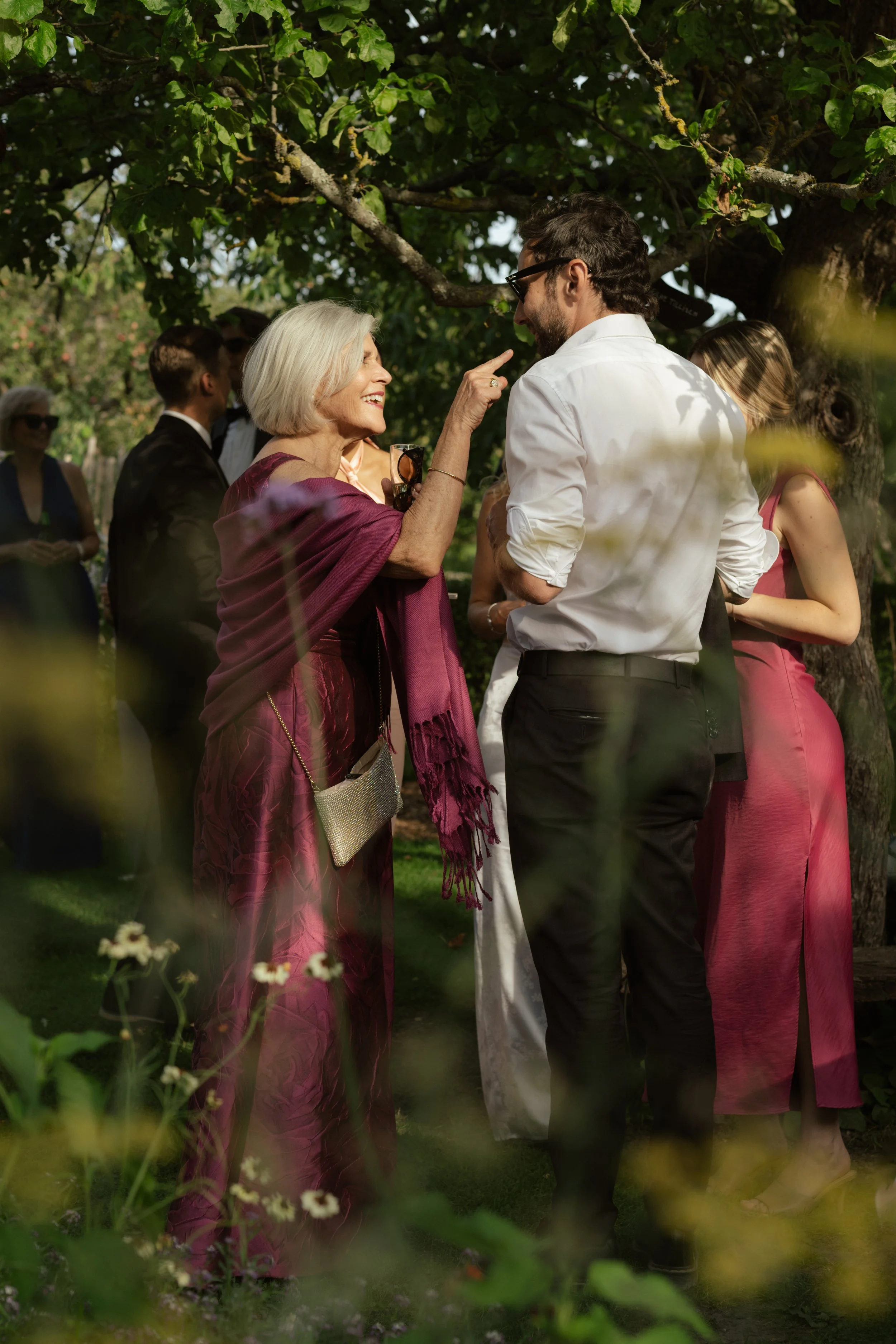 An elderly woman with gray hair and a young man with dark hair and glasses talking. The woman is smiling and pointing at the man, who is smiling back. They are outdoors under a tree, dressed in formal attire, with other people in the background.
