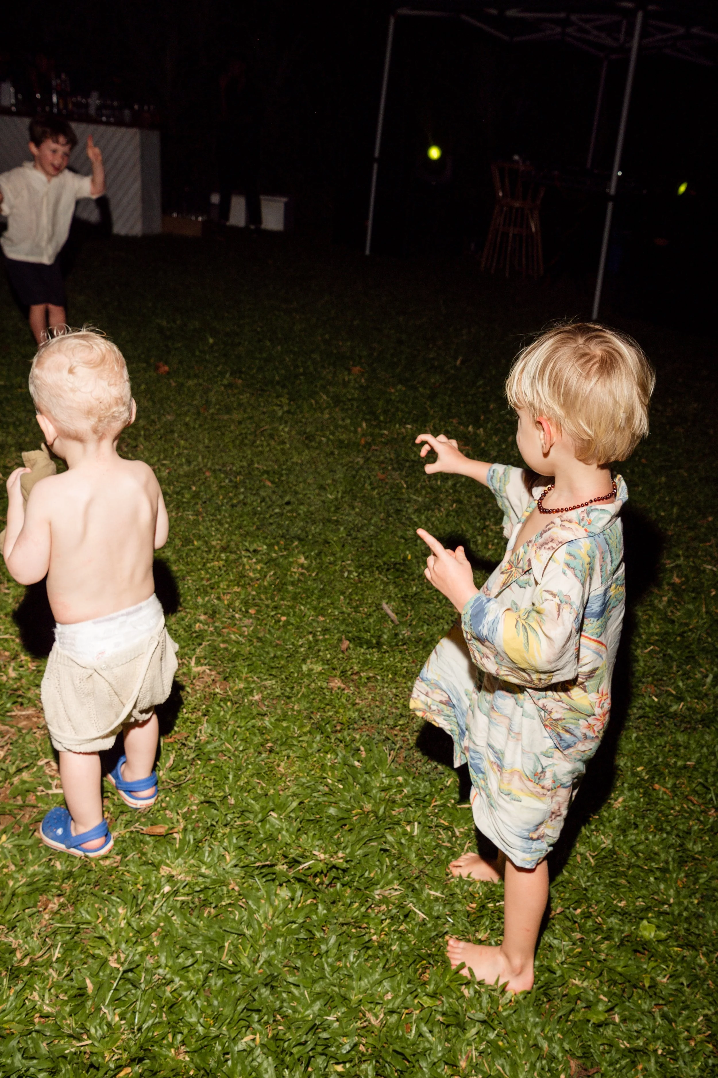 Children playing on a grassy yard at night, with one boy pointing and another boy in shorts and Crocs running away, girl in a colorful dress watching them.