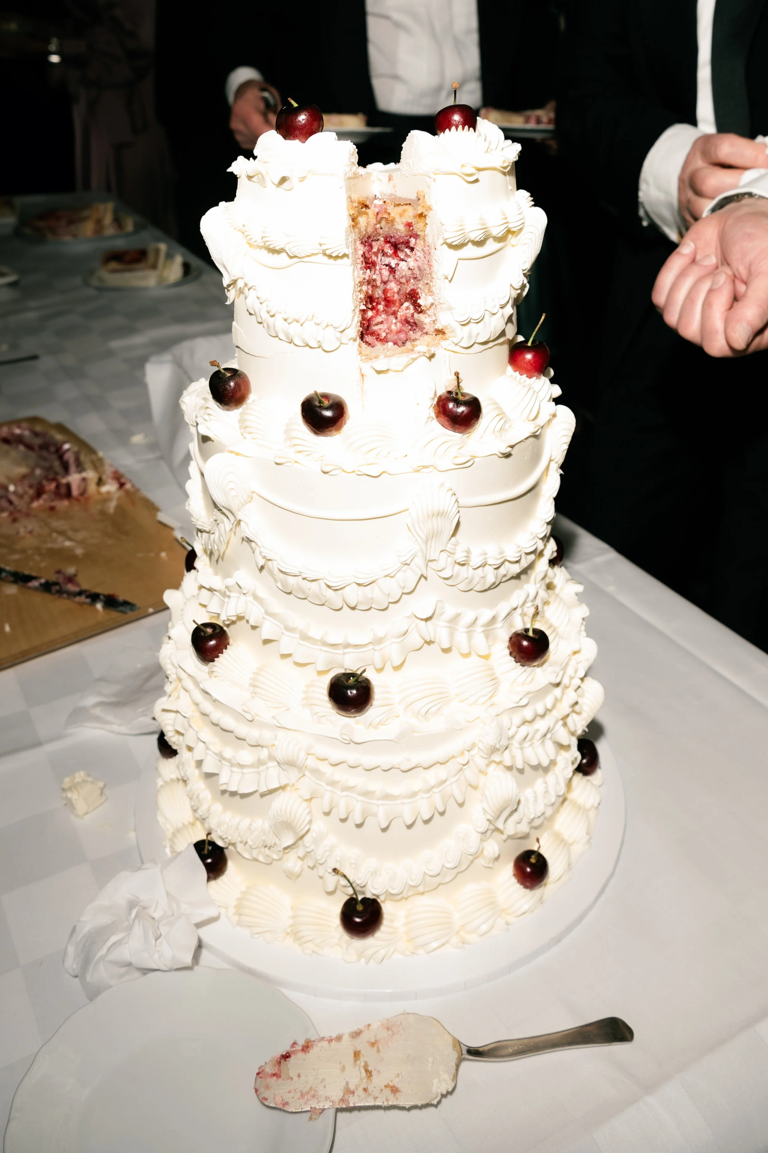 A tall, multi-layered white wedding cake decorated with cherries and ornate icing, with a slice removed showing pink and white interior layers.